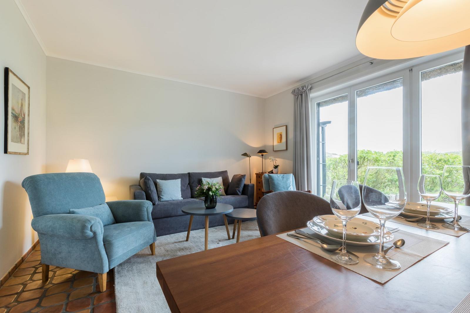 Wohn- und Essbereich mit Sofa, Sessel und Tisch. Große Fenster mit Blick auf Grün.