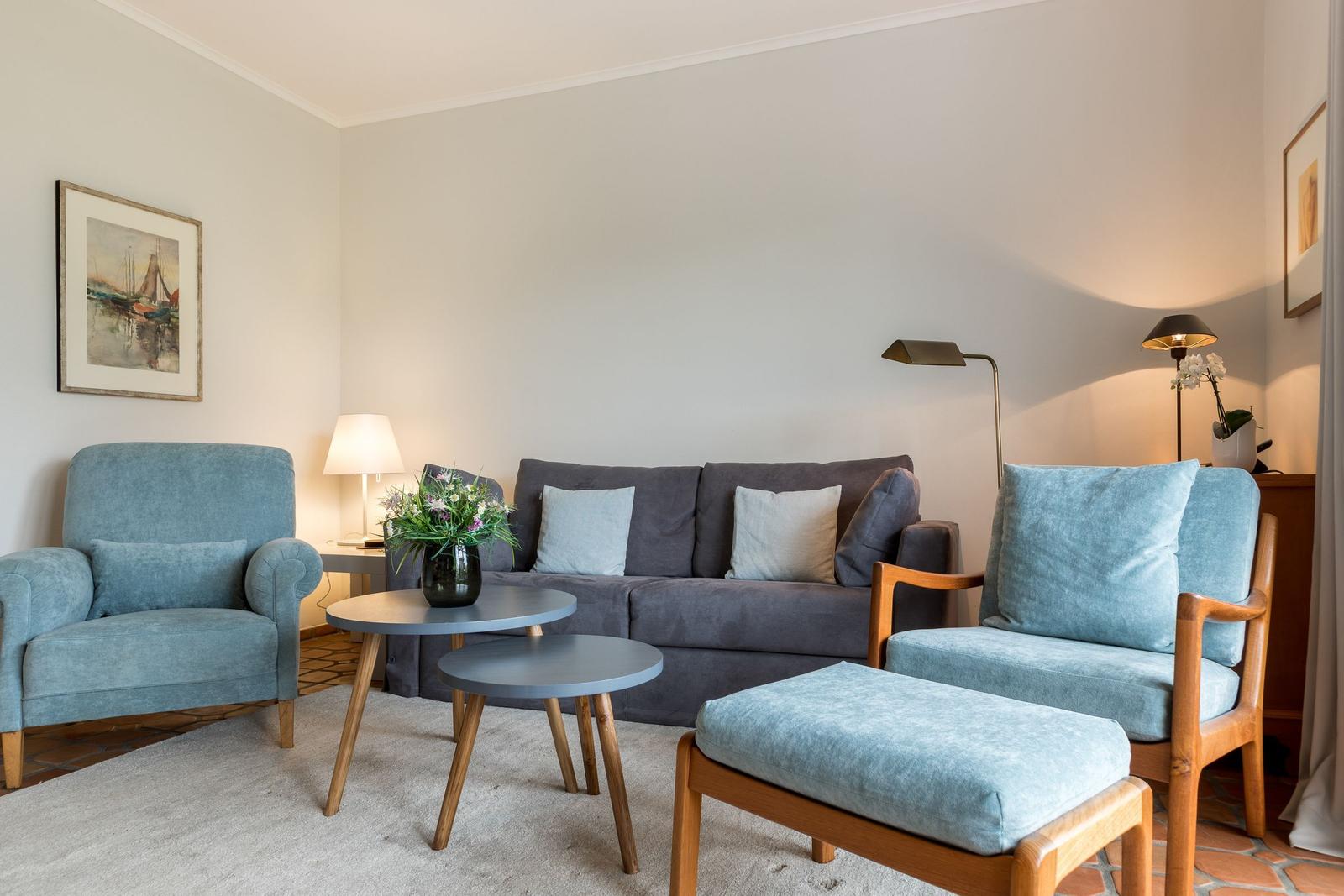 Wohnzimmer mit Sofa, zwei Sesseln und Tisch. Wandbild und Beleuchtung sichtbar.