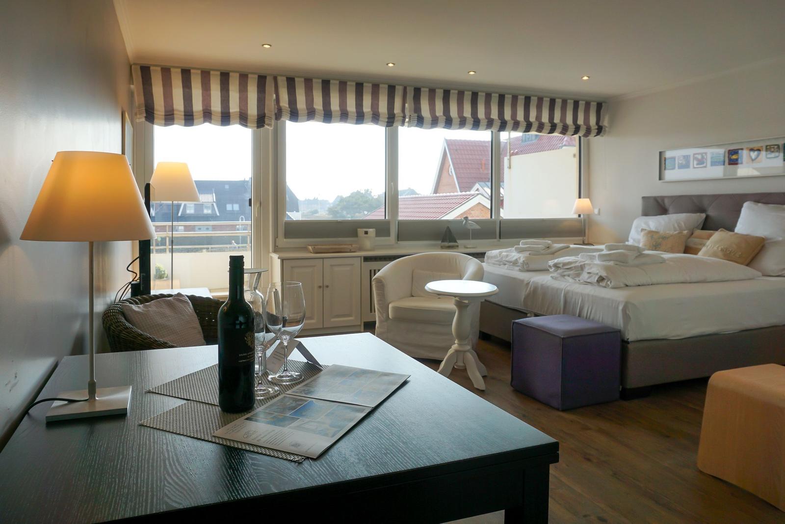 Schlafzimmer mit Bett, Sessel und Tisch. Fenster mit Streifenrollo. Holzboden und Lampen.