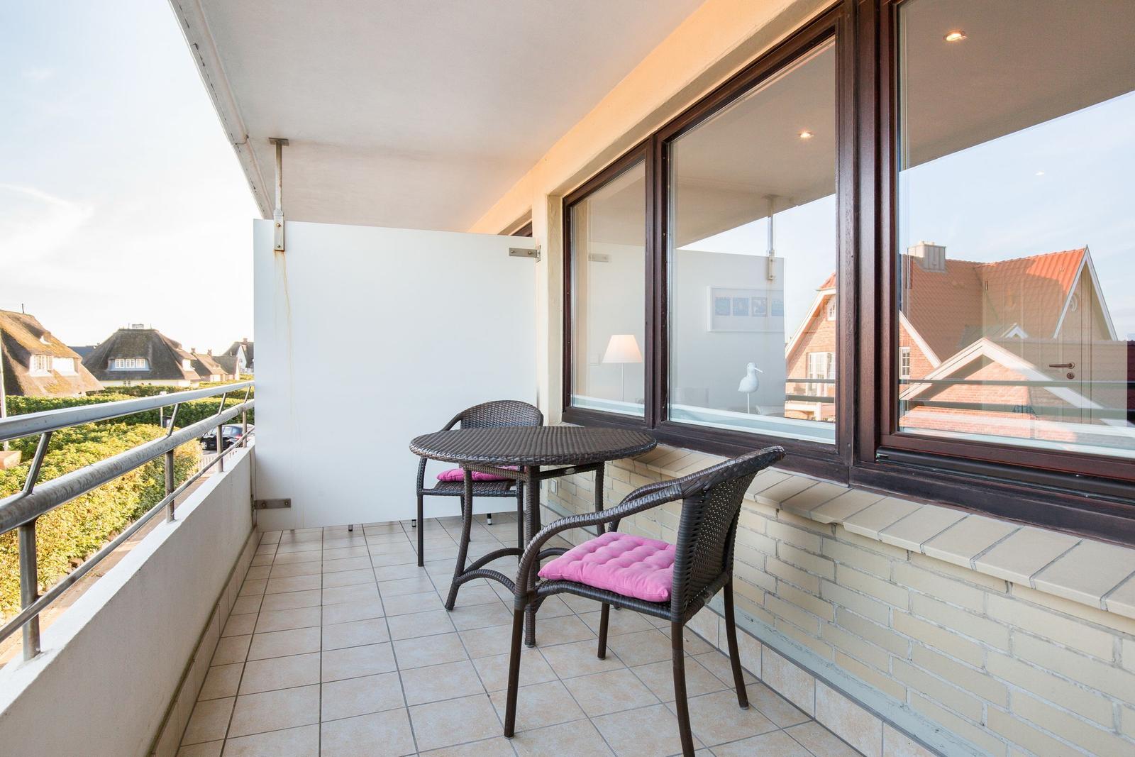 Terrasse mit Tisch und zwei Stühlen mit pinken Kissen. Blick auf Dächer und Grünflächen.