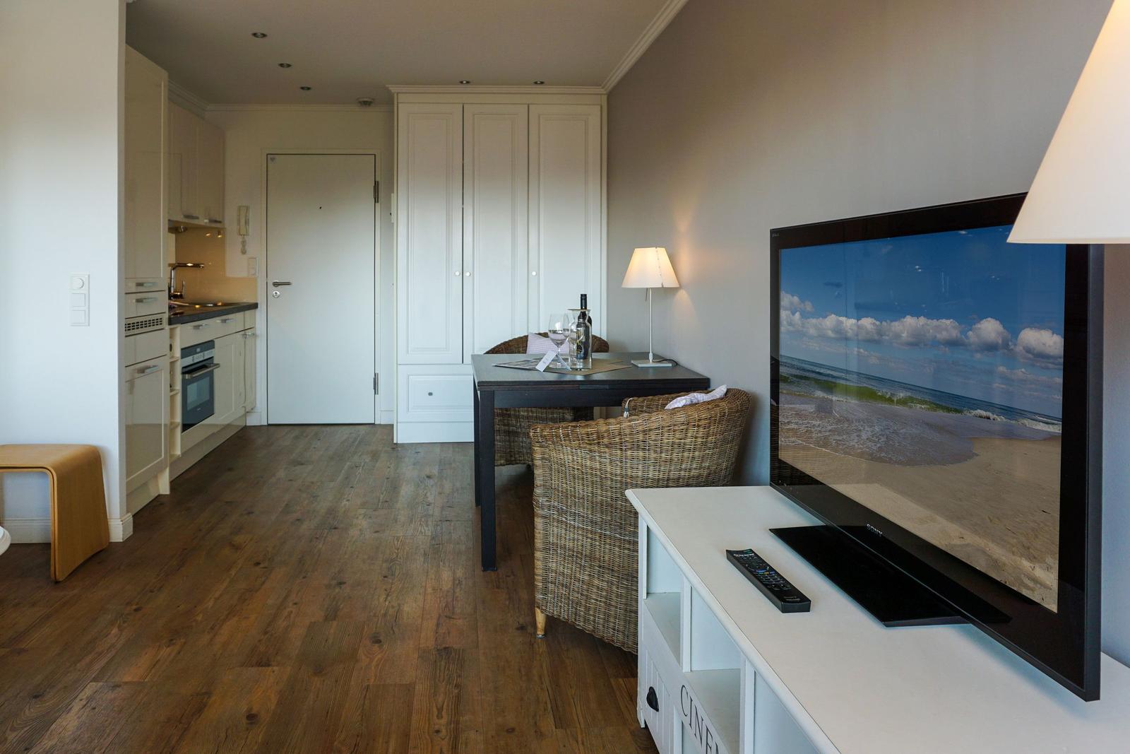 Wohnbereich mit TV, Esstisch und Küchenbereich. Holzfußboden und weiße Möbel.