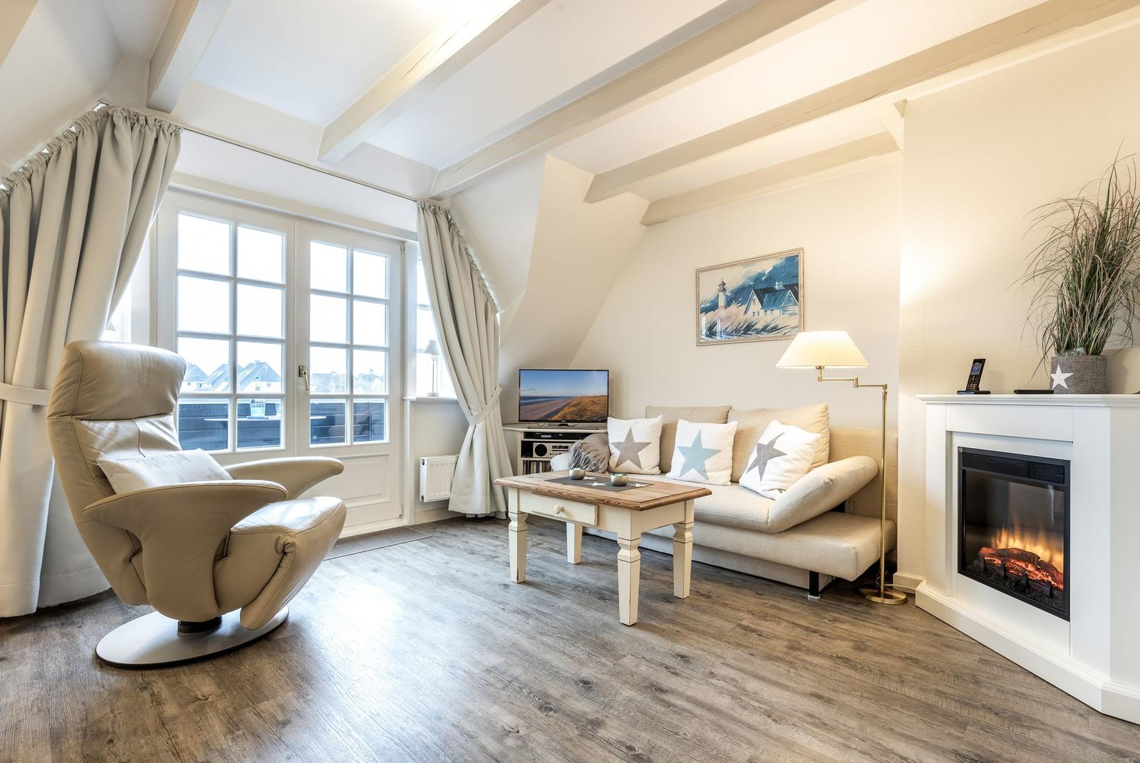 Wohnzimmer mit Sessel, Sofa, Kamin und TV. Holzboden und große Fenster.