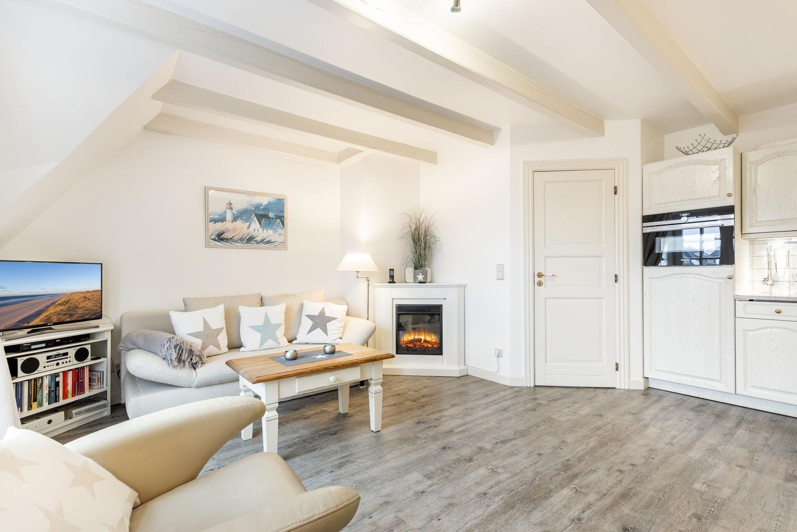 Wohnbereich mit Sofa, Kamin und Küchenzeile. Holzboden und weiße Einrichtung.