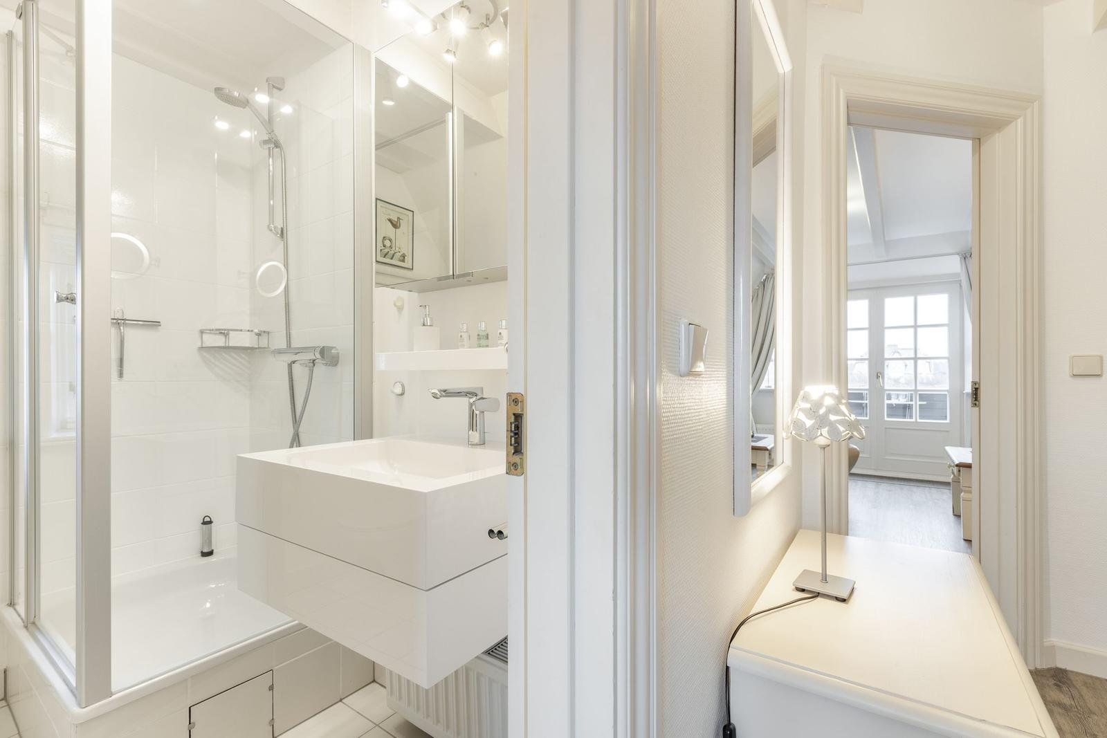 Badezimmer mit Dusche, Waschbecken und Spiegel. Blick in angrenzenden Raum.