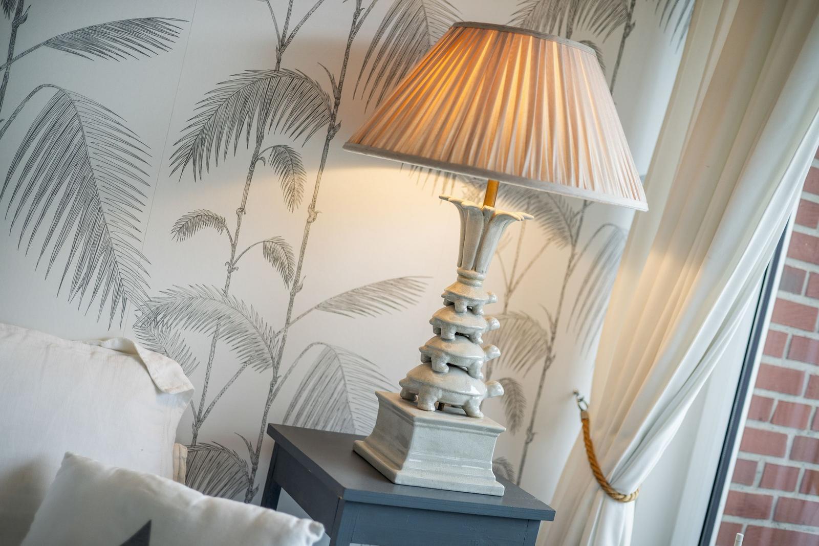 Table lamp on nightstand beside bed, wall with palm pattern, white curtains.