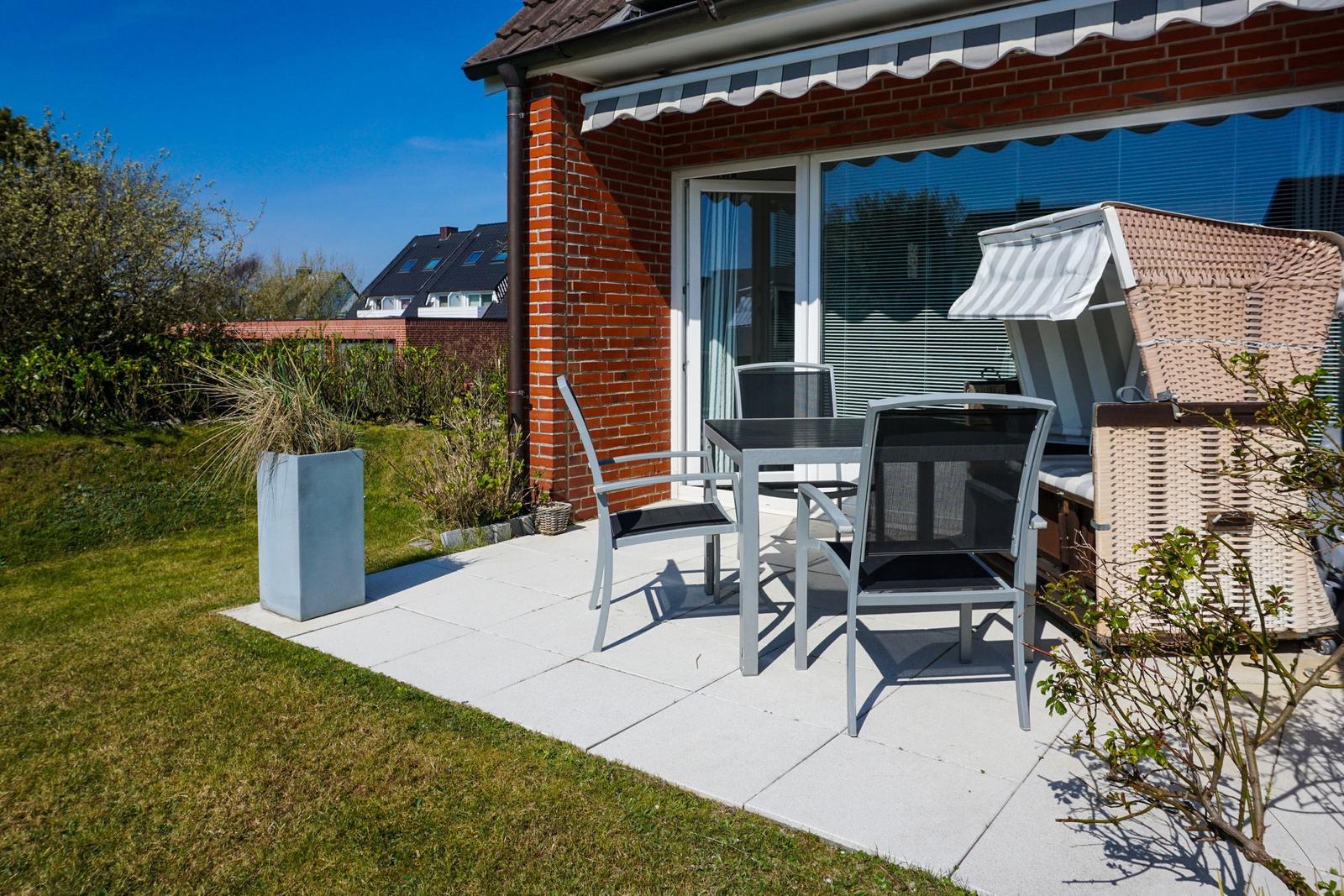 Terrace with table, chairs, and lounge under awning beside red brick house.