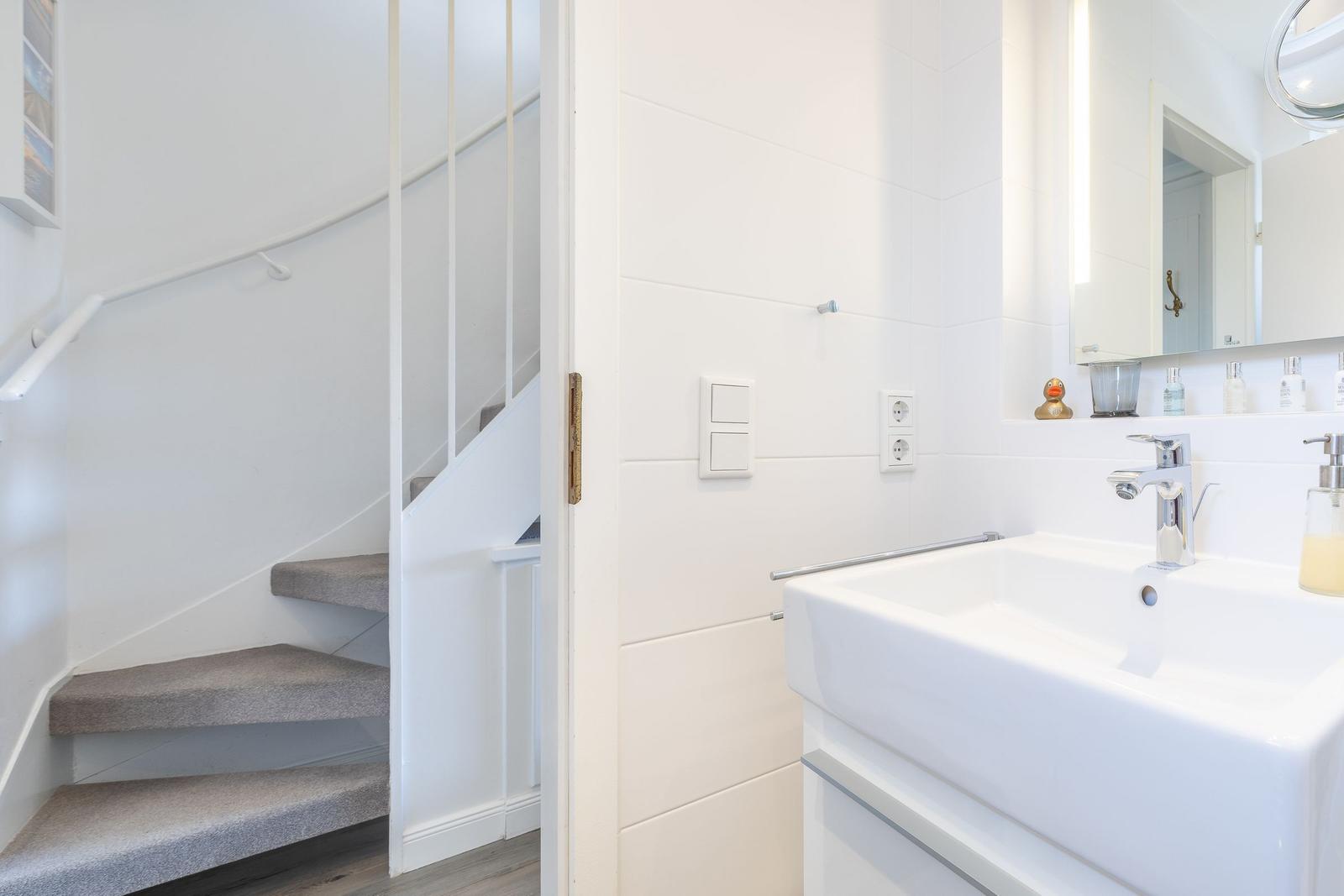 Badezimmer mit Waschbecken und Spiegel. Treppenhaus im Hintergrund.