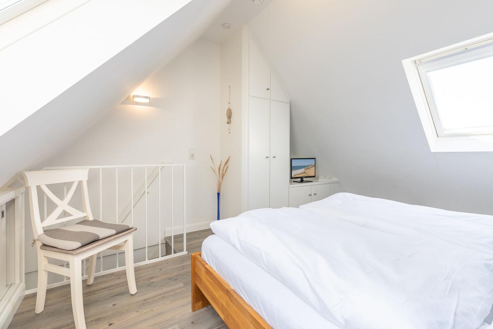 Schlafzimmer mit Dachschrägen, weißer Bettwäsche und Holzbett. Fenster und Stuhl sichtbar.