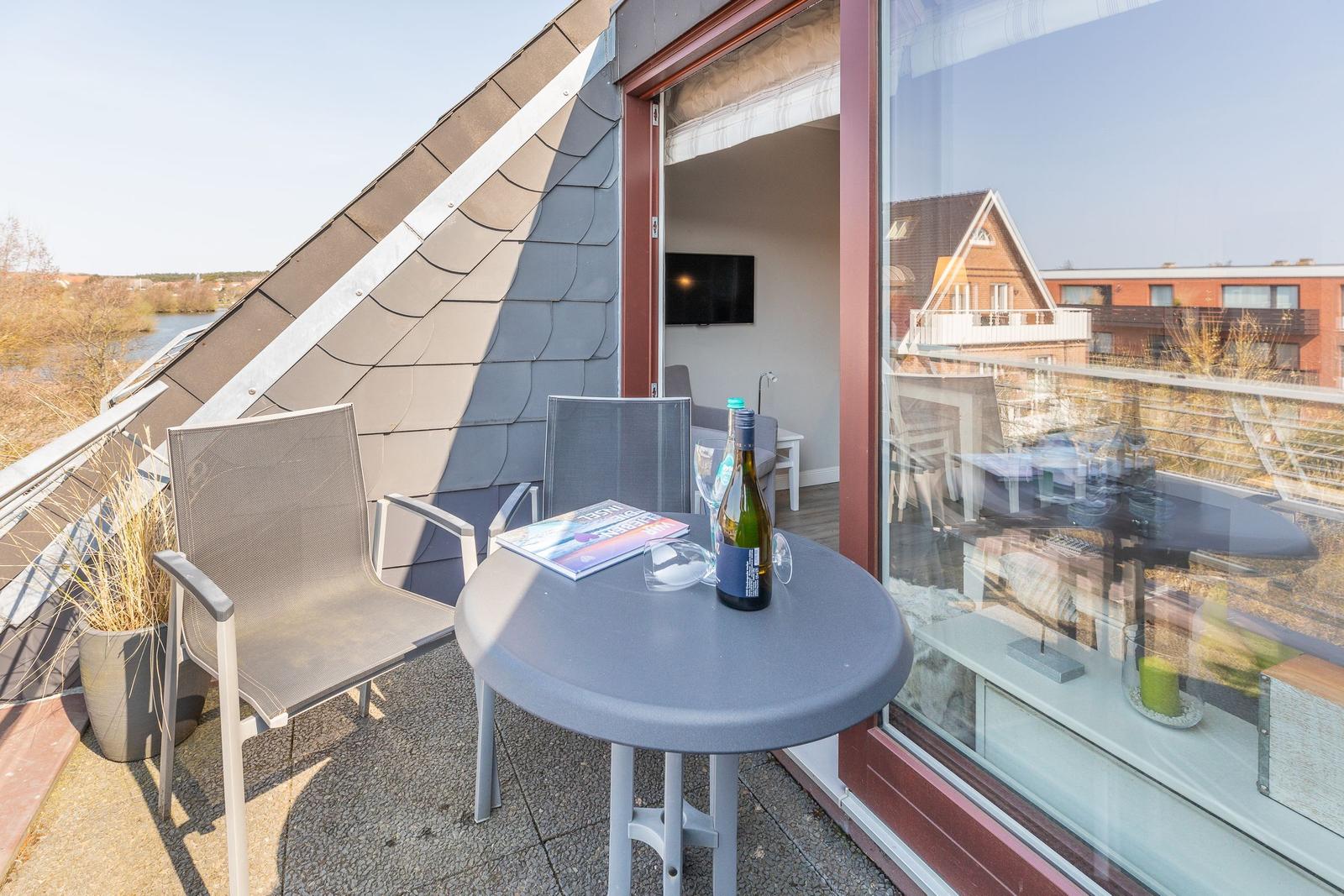 Terrasse mit Tisch, zwei Stühlen und Flasche. Blick auf Wasser und Gebäude.