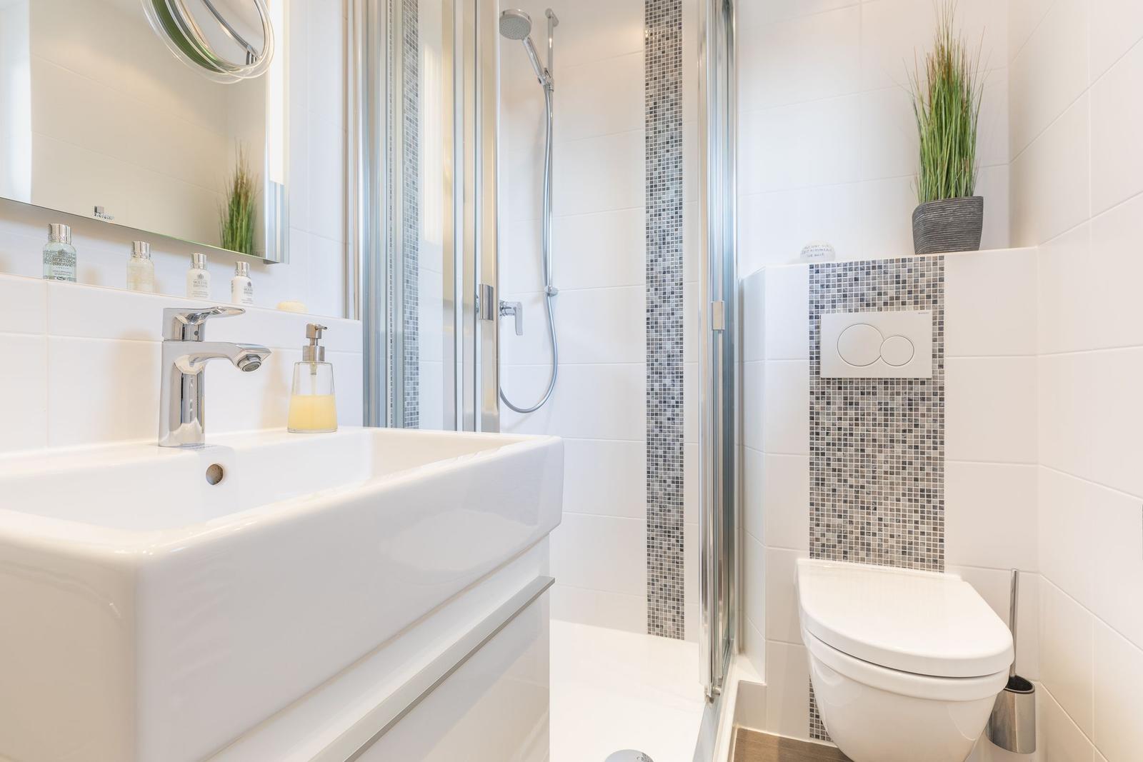 Badezimmer mit Dusche, Waschbecken und Toilette. Weiße Fliesen und Mosaikverkleidung.