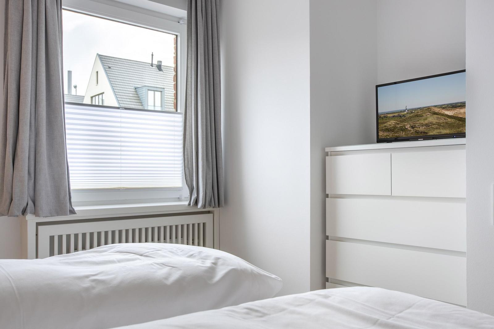 Bedroom with white bed, window with blinds, and TV on dresser.