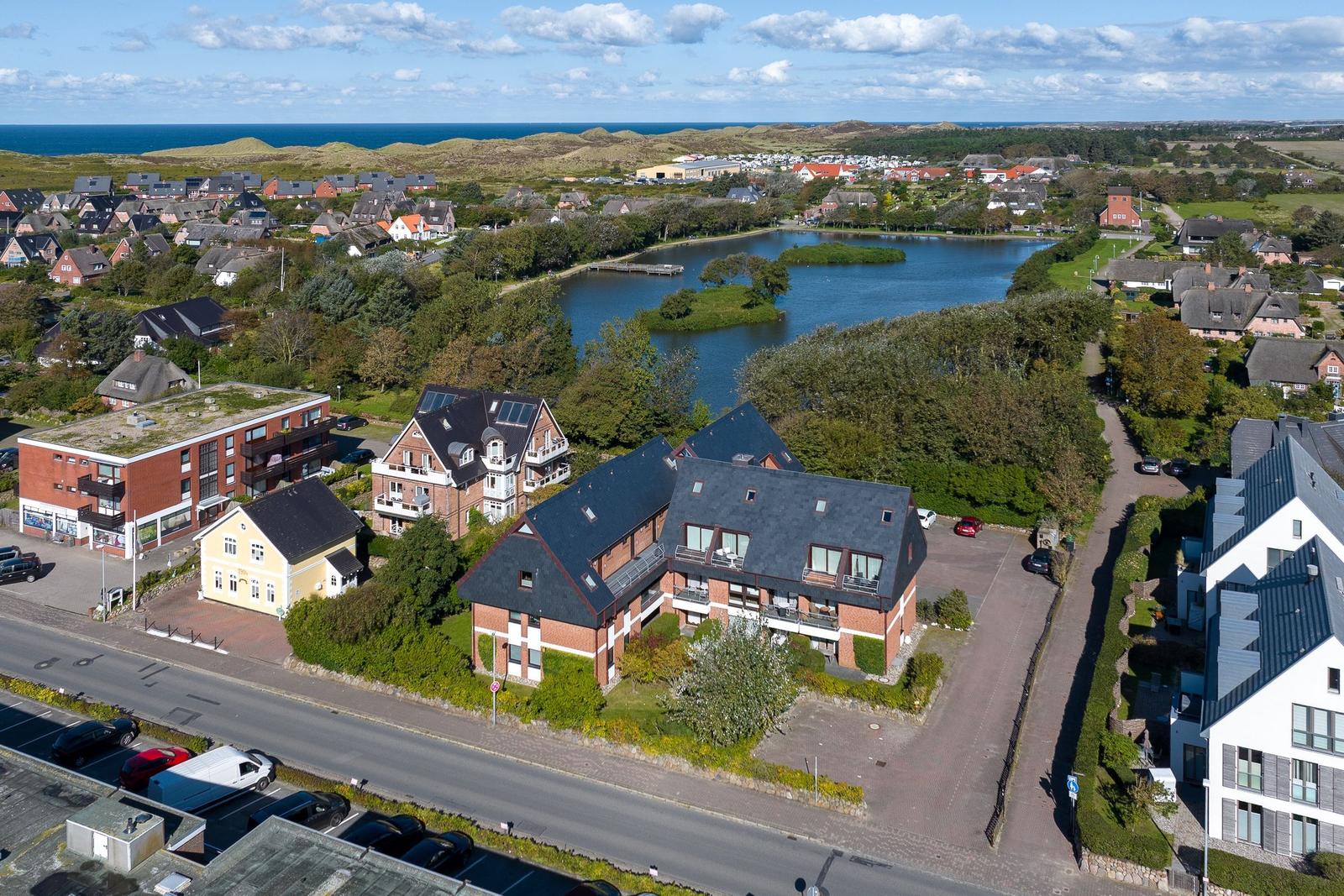 House with balcony, surrounded by greenery and a lake. Houses and the sea are visible in the background.