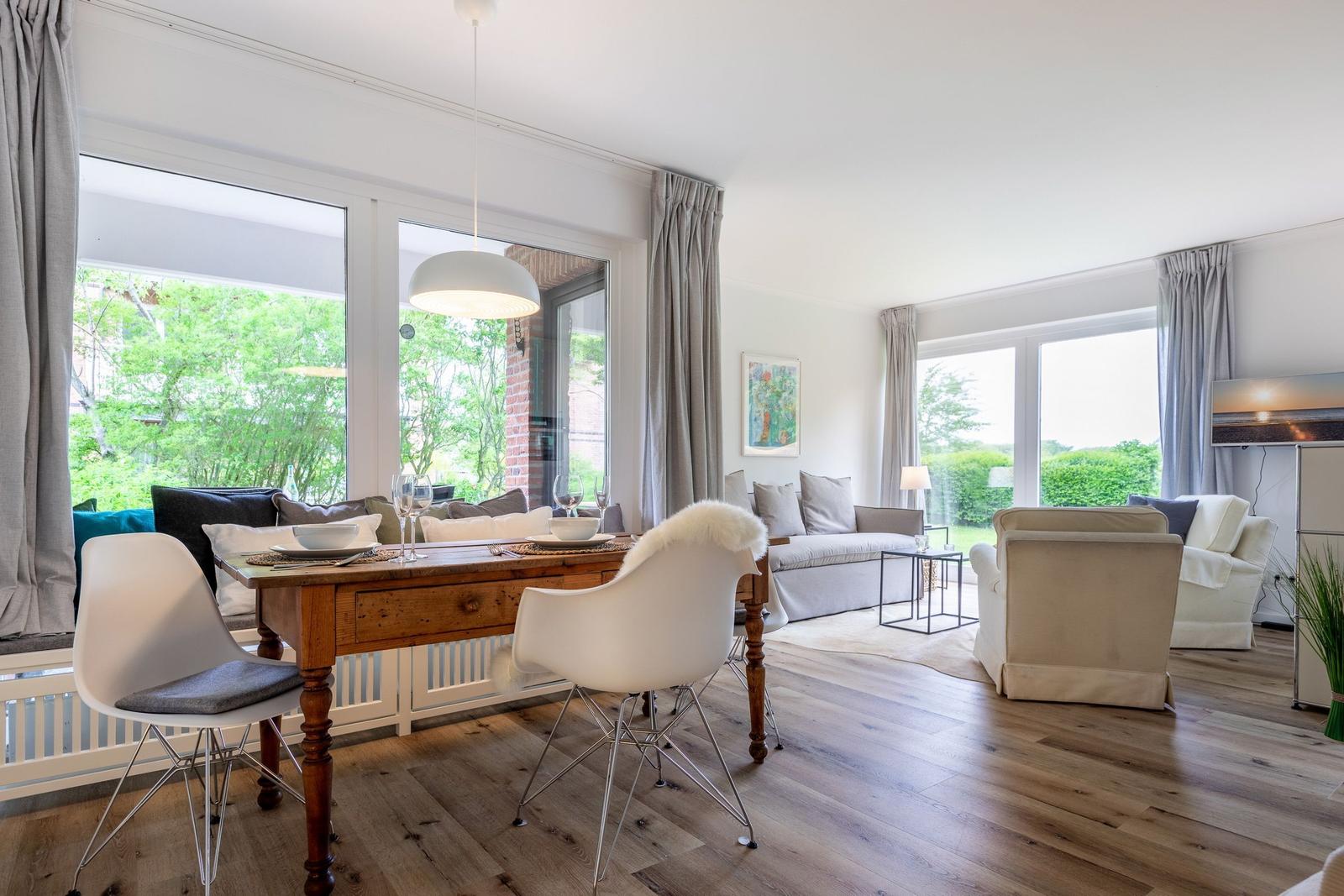Dining area with wooden table and white chairs. Living area with sofa and armchairs. Large windows with garden view.
