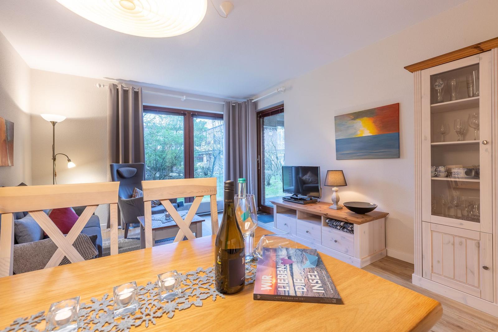 Dining area with wooden table, wine bottle, and book. Background: living room with TV and glass cabinet.