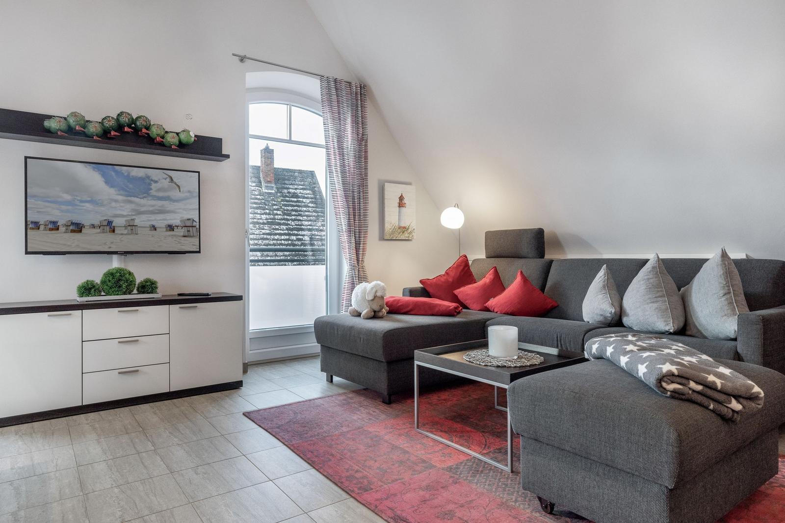 Wohnzimmer mit Sofa, Fernseher und Fenster. Auf dem Sofa liegen Kissen und eine Decke.