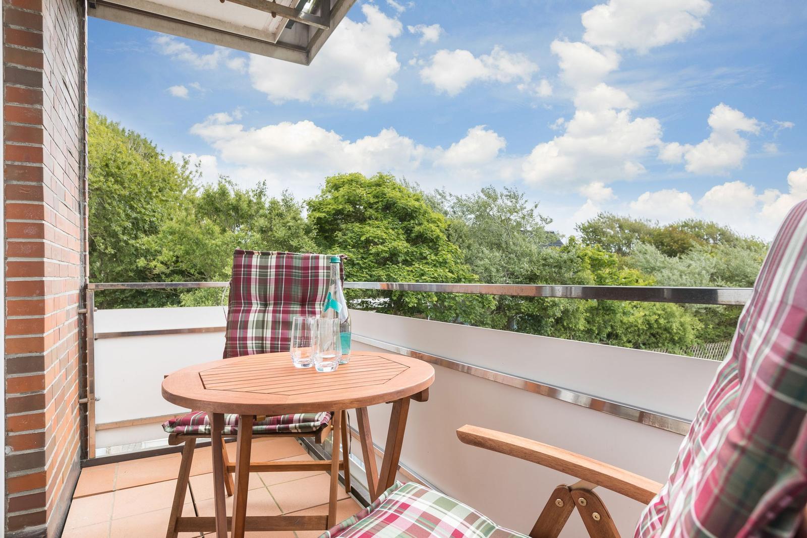 Balkon mit Tisch, zwei Stühlen und Blick auf Bäume unter blauem Himmel.