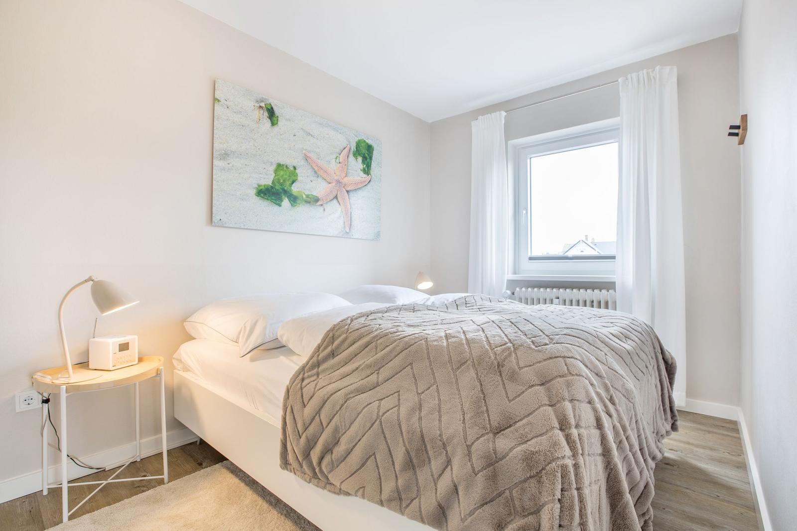 Schlafzimmer mit Bett, Wandbild und Fenster mit weißen Vorhängen.