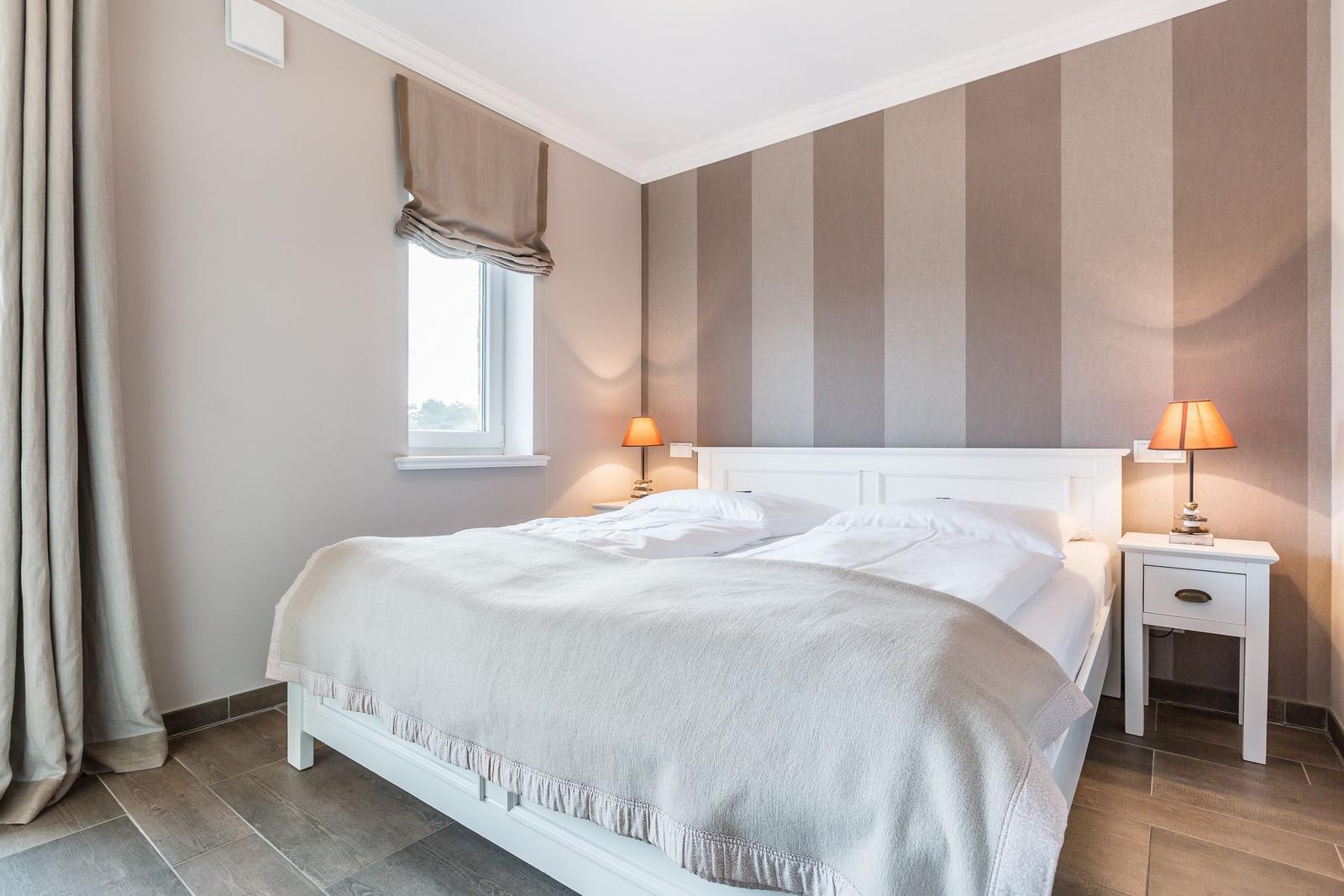 Schlafzimmer mit Bett, Nachttisch und Lampe. Wand mit Streifenmuster. Fenster mit Rollladen.