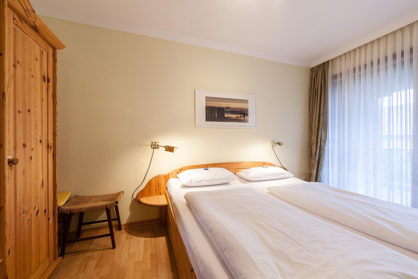 Doppelbett mit weißen Laken, Holzkommode und Wandbild. Fenster mit Vorhängen.