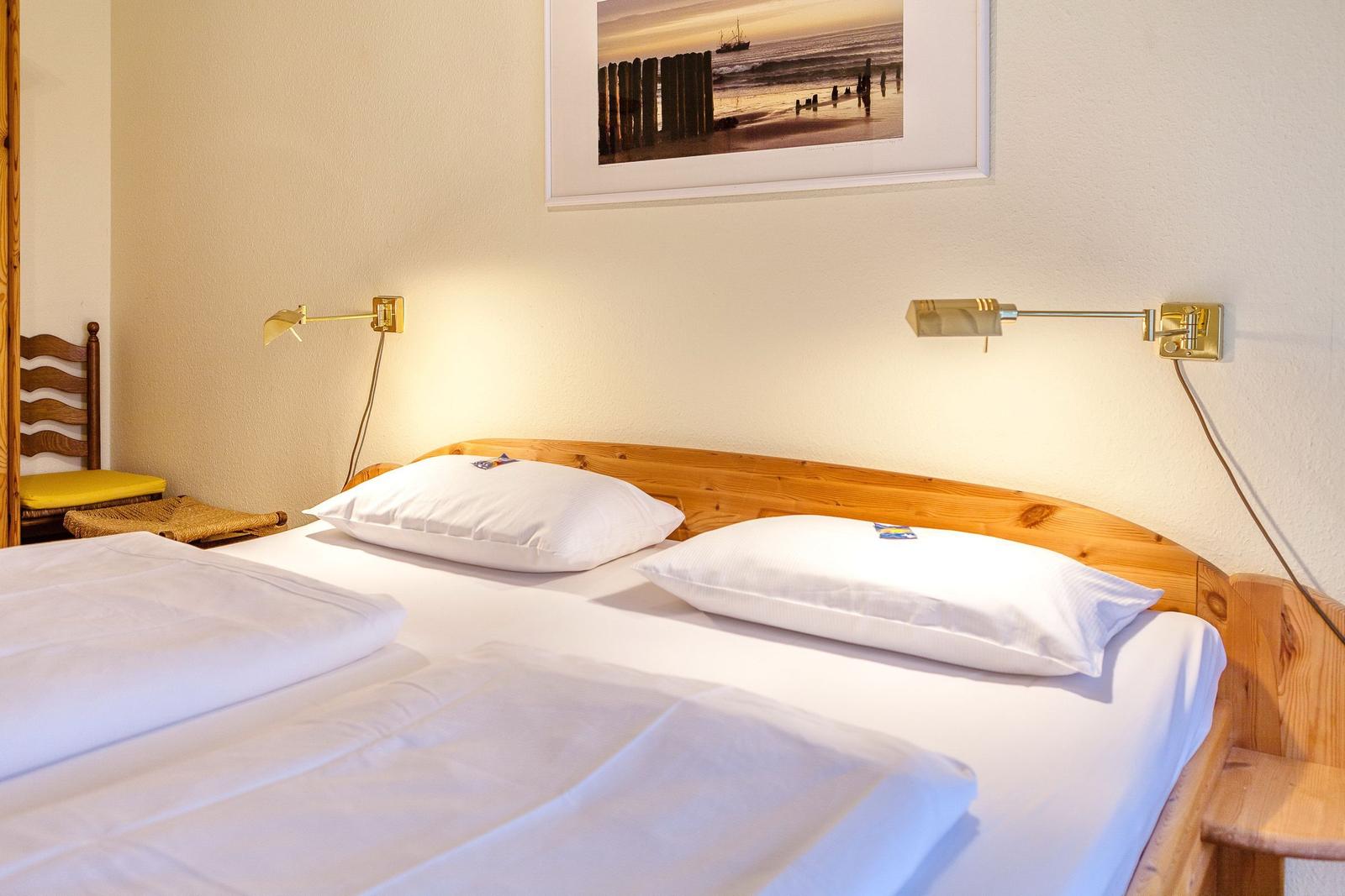 Ein Bett mit weißer Bettwäsche und Holzrahmen. An der Wand hängt ein Bild und zwei Wandlampen.