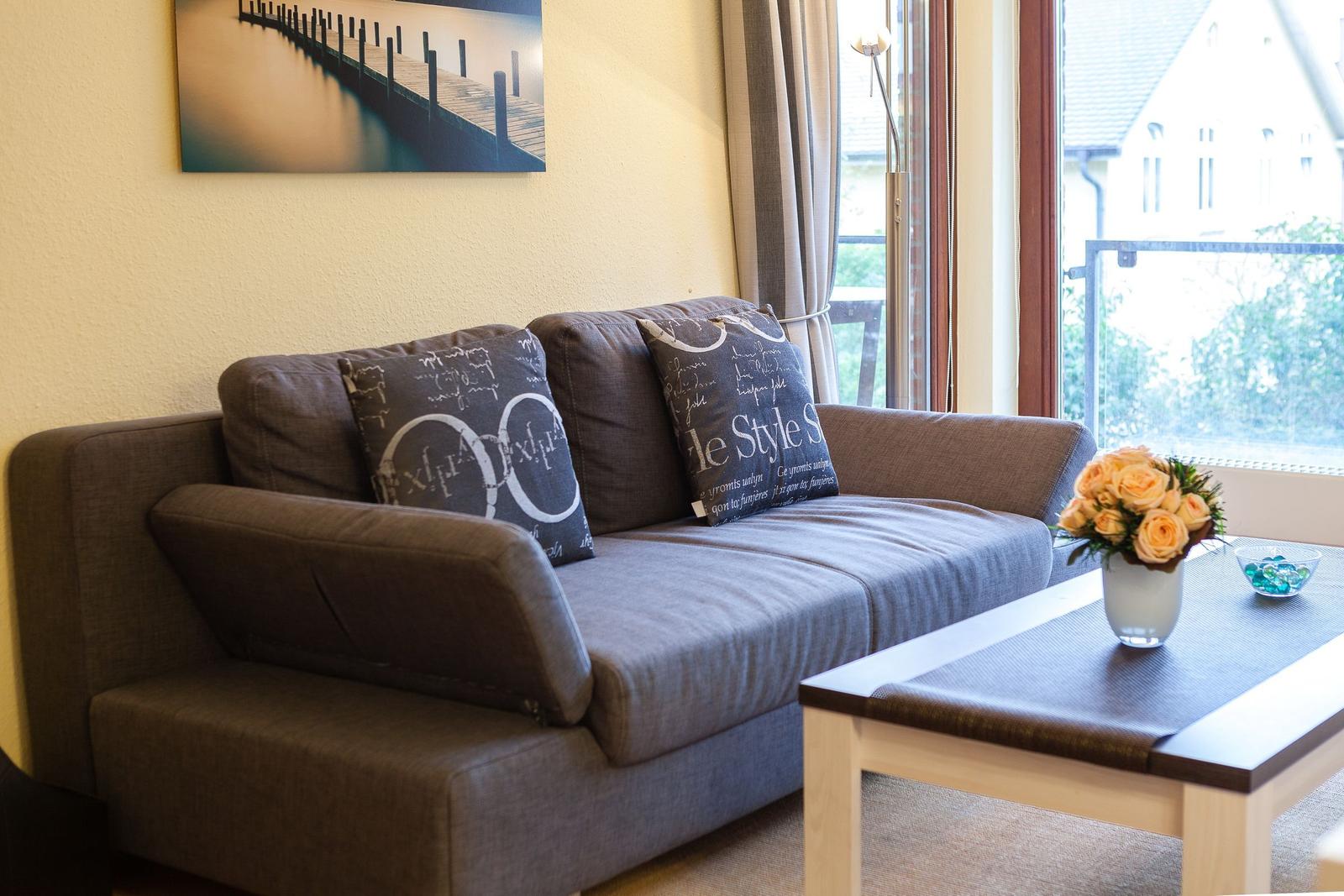 Ein Sofa mit Kissen steht vor einem Fenster. Auf dem Tisch stehen Blumen und eine Schale.