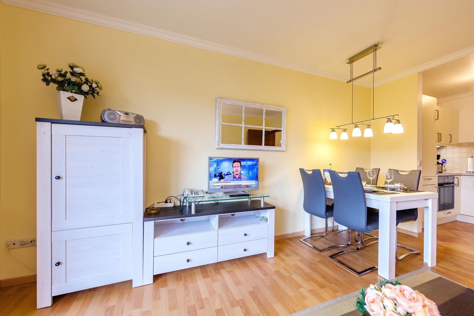 Wohn- und Essbereich mit Fernseher, Tisch und Stühlen sowie Küchenbereich im Hintergrund.