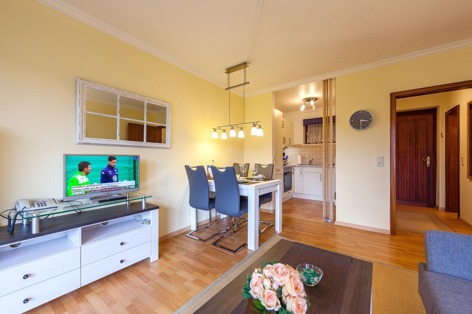 Wohn- und Essbereich mit TV, Tisch und Stühlen, Küche im Hintergrund.