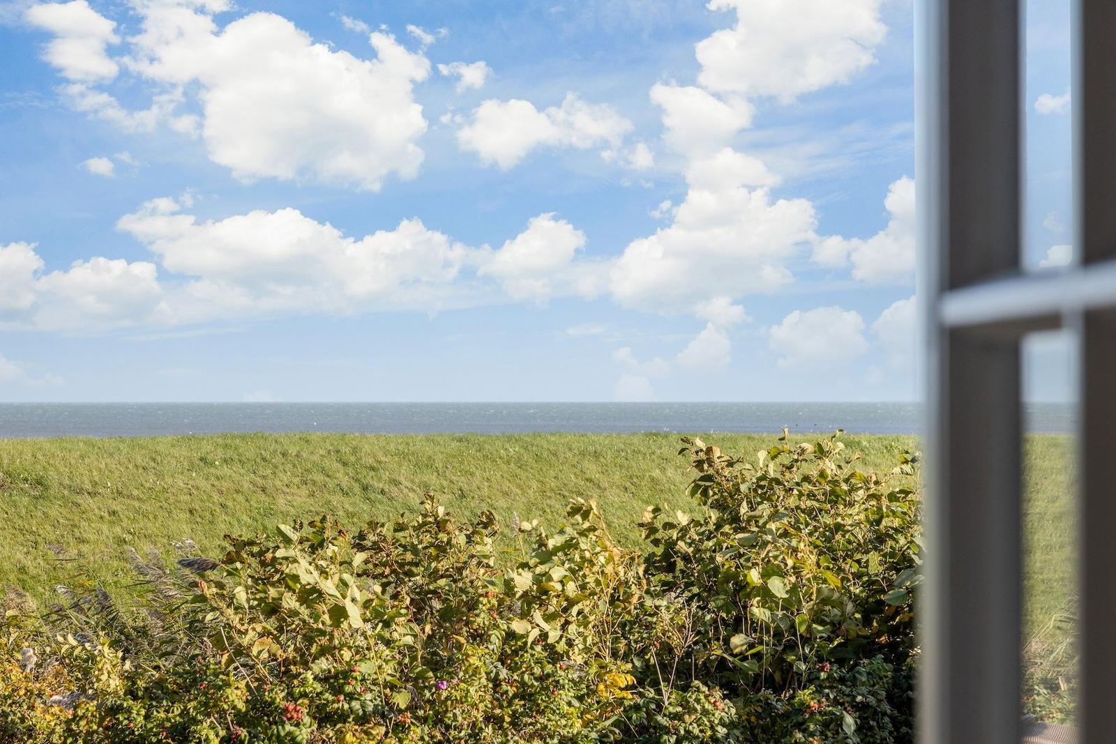 Durch ein Fenster blickt man auf einen grünen Hang und das Meer unter einem blauen Himmel mit weißen Wolken.