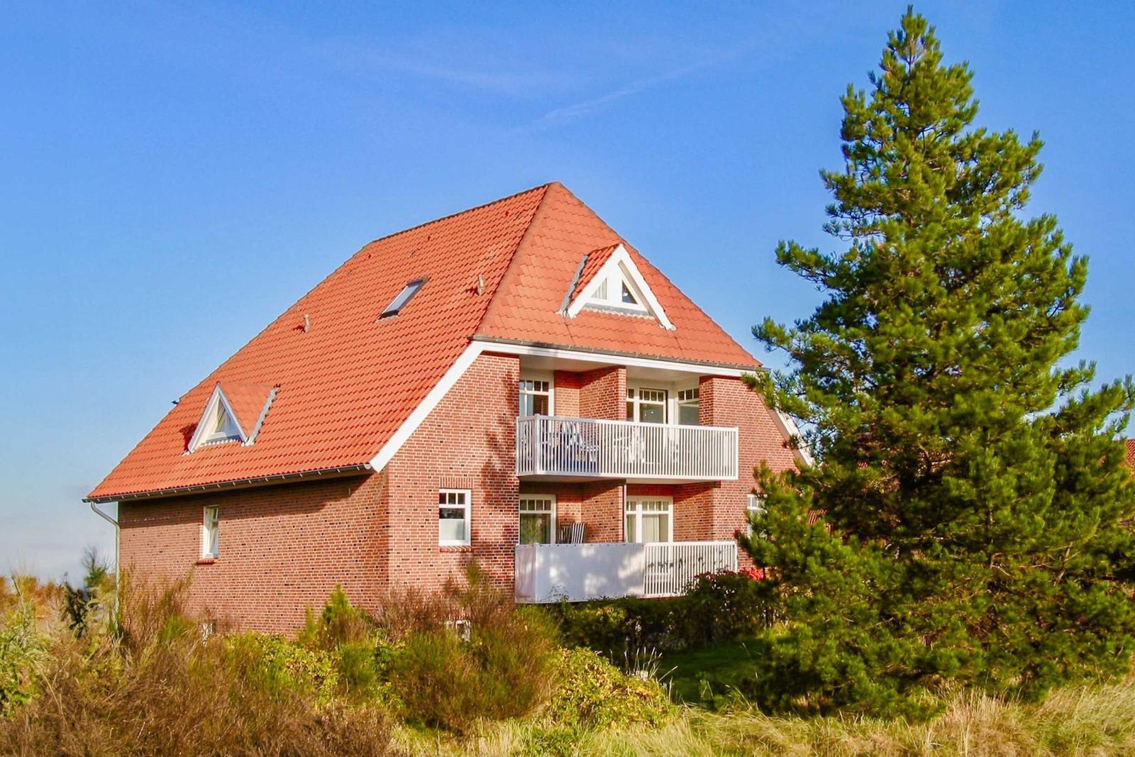 Ein zweistöckiges Haus mit rotem Dach und weißen Balkonen. Umgeben von Gras und einem großen Baum.