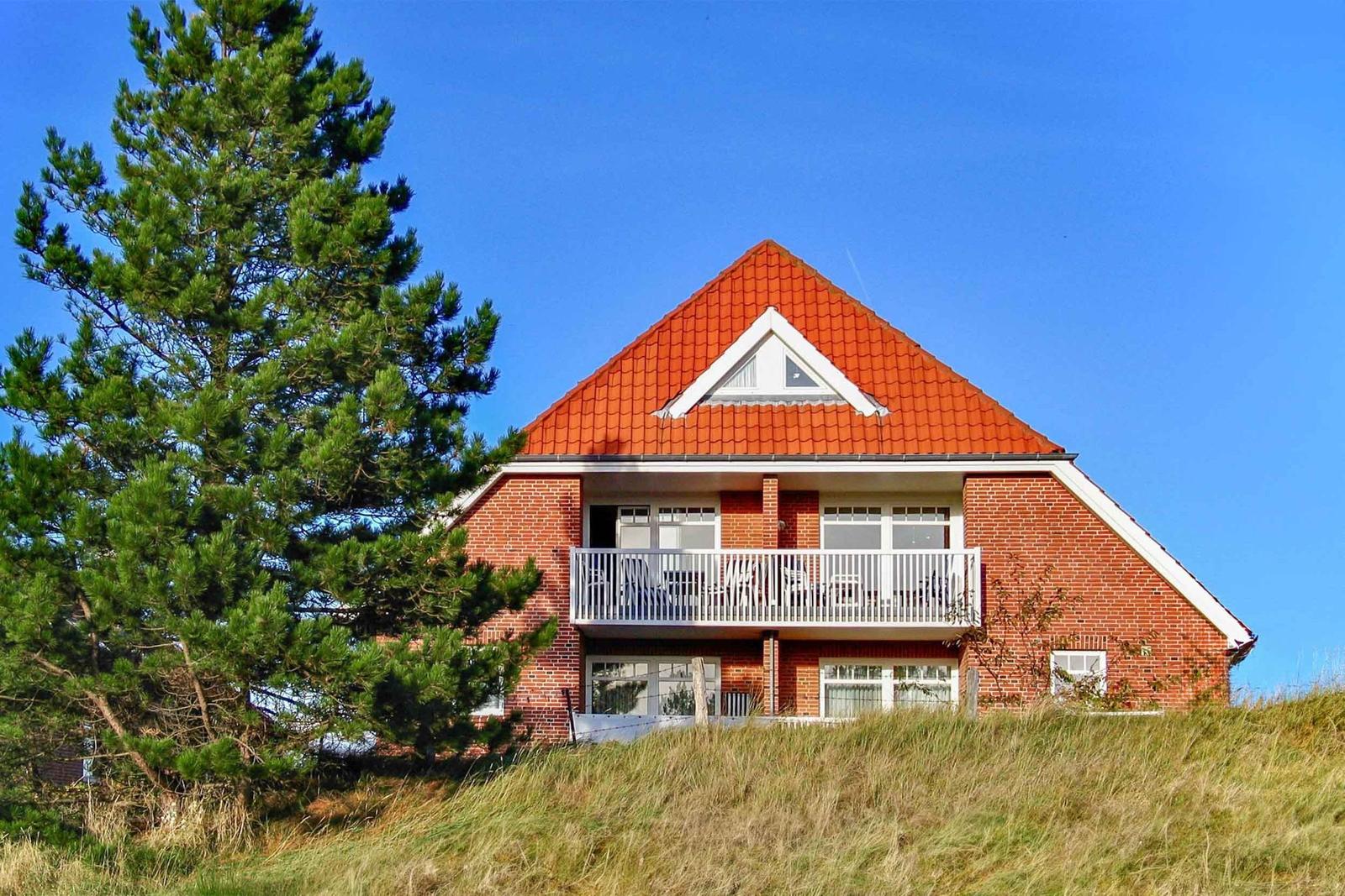 Zweistöckiges Haus mit Balkon, rotem Dach und Backsteinfassade auf grüner Anhöhe.