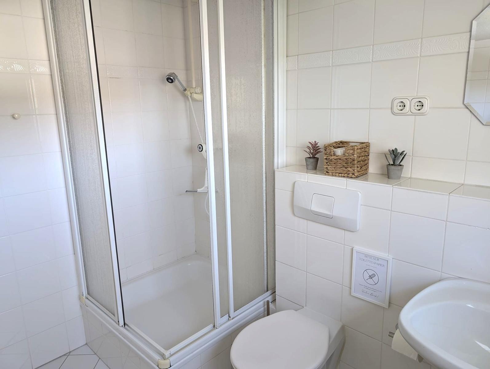 Badezimmer mit Dusche, Toilette und Waschbecken. Weiße Fliesen und kleine Pflanzen.