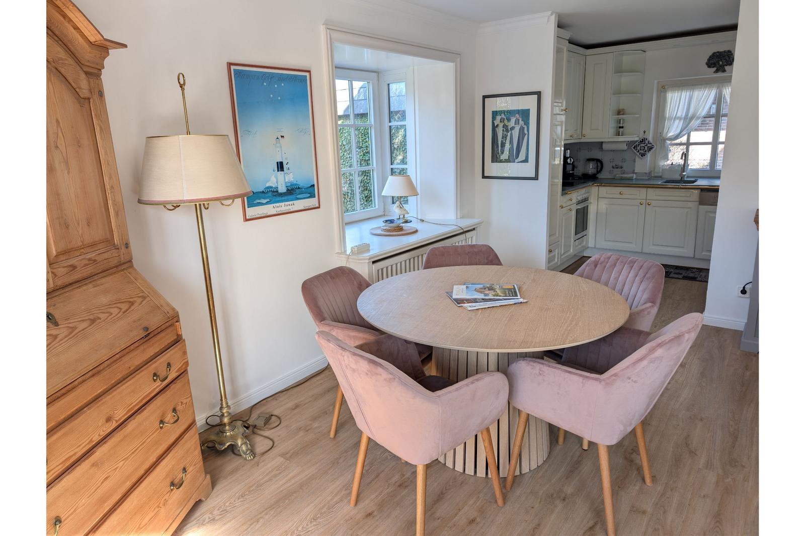 Round table with four pink chairs, beside wooden cabinet and lamps. Kitchen in background.
