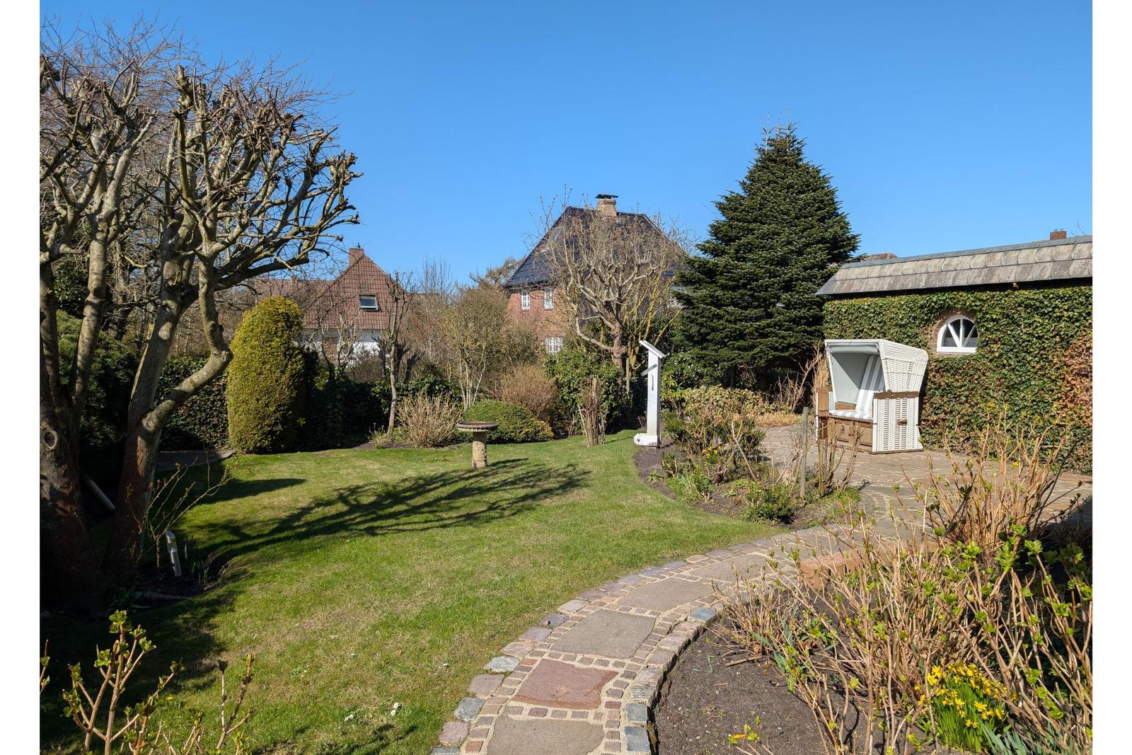 Garden with stone column, paths, and pavilion. Background: buildings and trees.