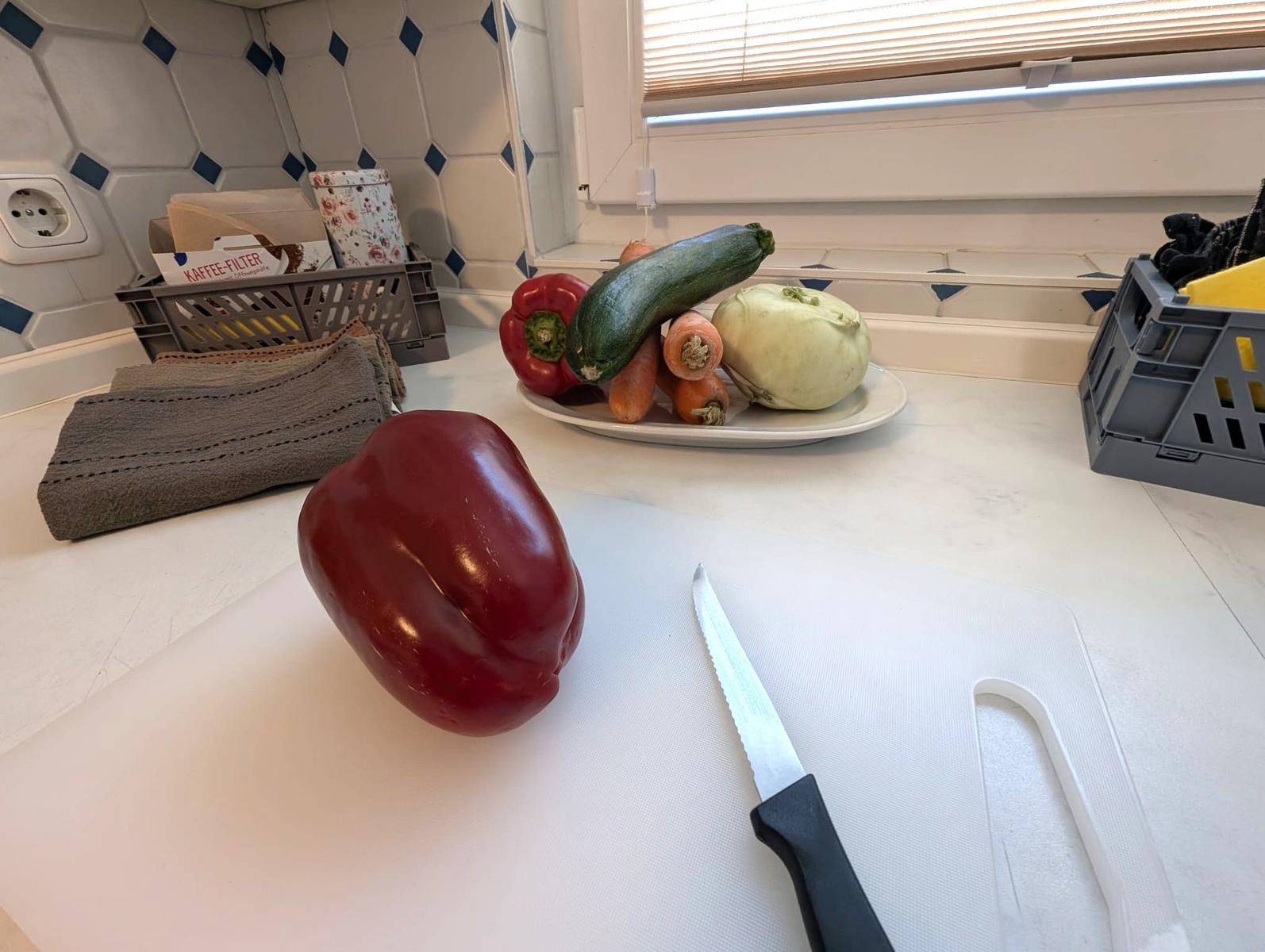 Küchenarbeitsfläche mit rotem Paprika, Messer und Gemüse auf Teller.