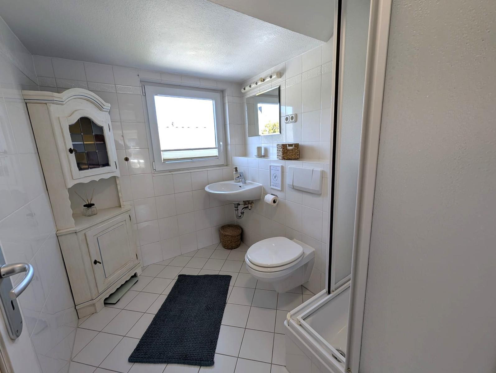 Badezimmer mit Toilette, Waschbecken und Dusche. Weiße Fliesen und ein dunkler Teppich.