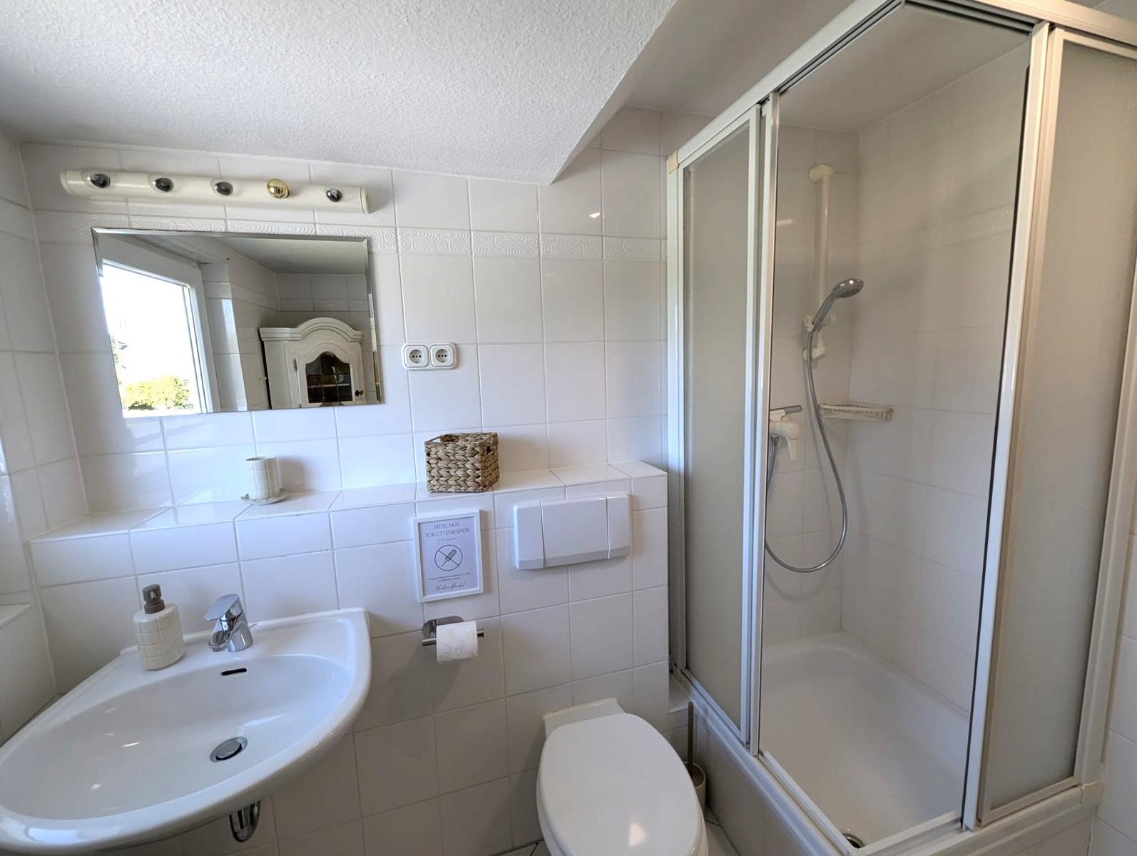 Badezimmer mit Dusche, Waschbecken und Toilette. Weiße Fliesen und Spiegel.