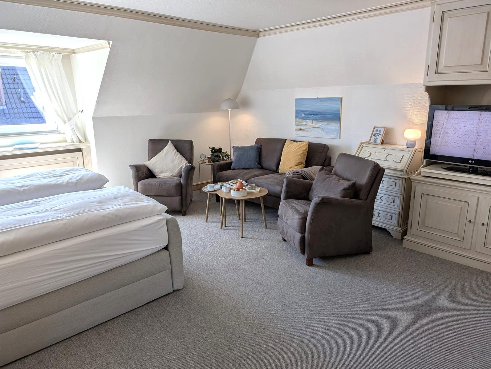 Schlafzimmer mit Sofa, zwei Sesseln und Fernseher. Fenster mit Blick auf Dach.