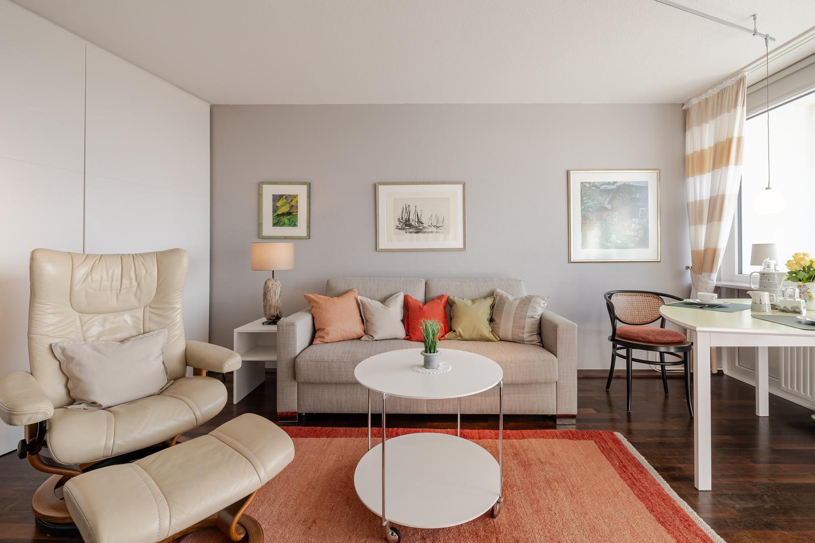Wohnzimmer mit Sofa, Sessel und Esstisch. Wandbilder und Fenster mit Vorhängen.
