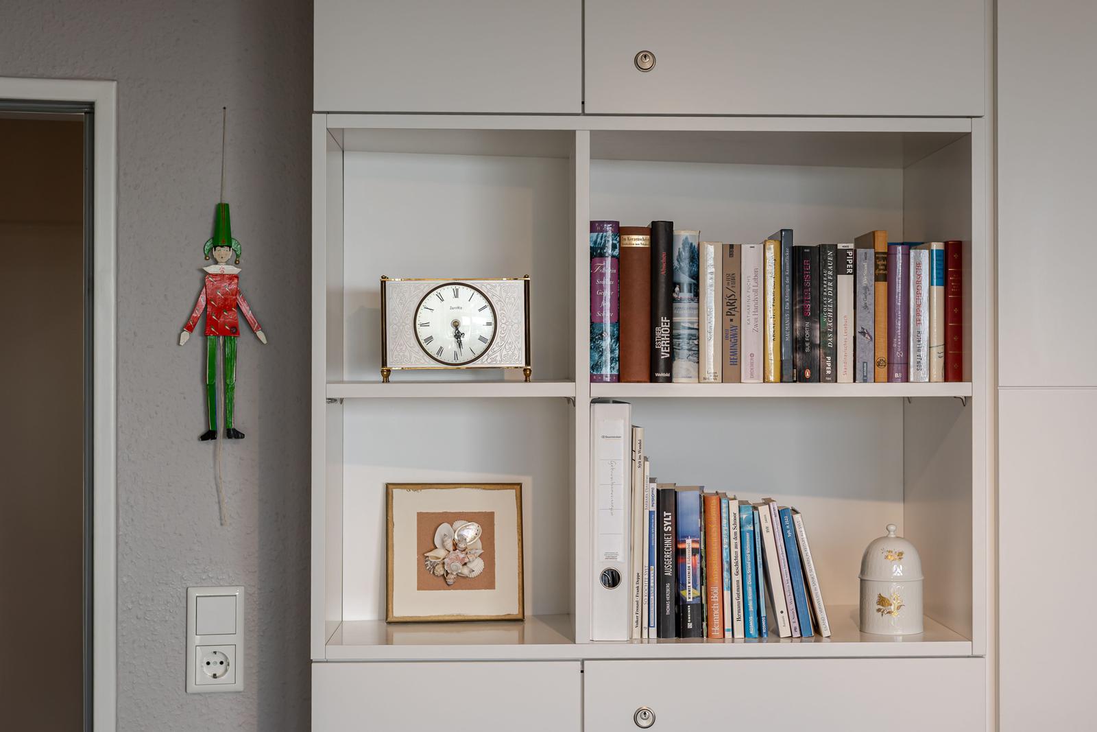 Bücherregal mit Uhr, Bilderrahmen und Dekoration. Wandhängendes Figurenobjekt neben Tür.