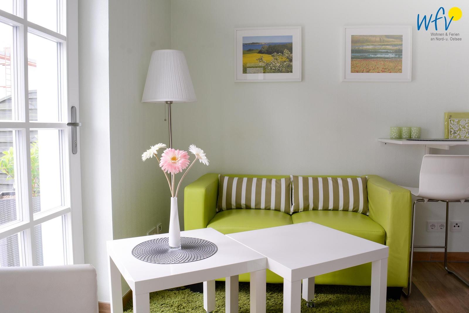 Ein Wohnzimmer mit grünem Sofa, weißen Tischen und Fenster. Wandbilder und Stehlampe.