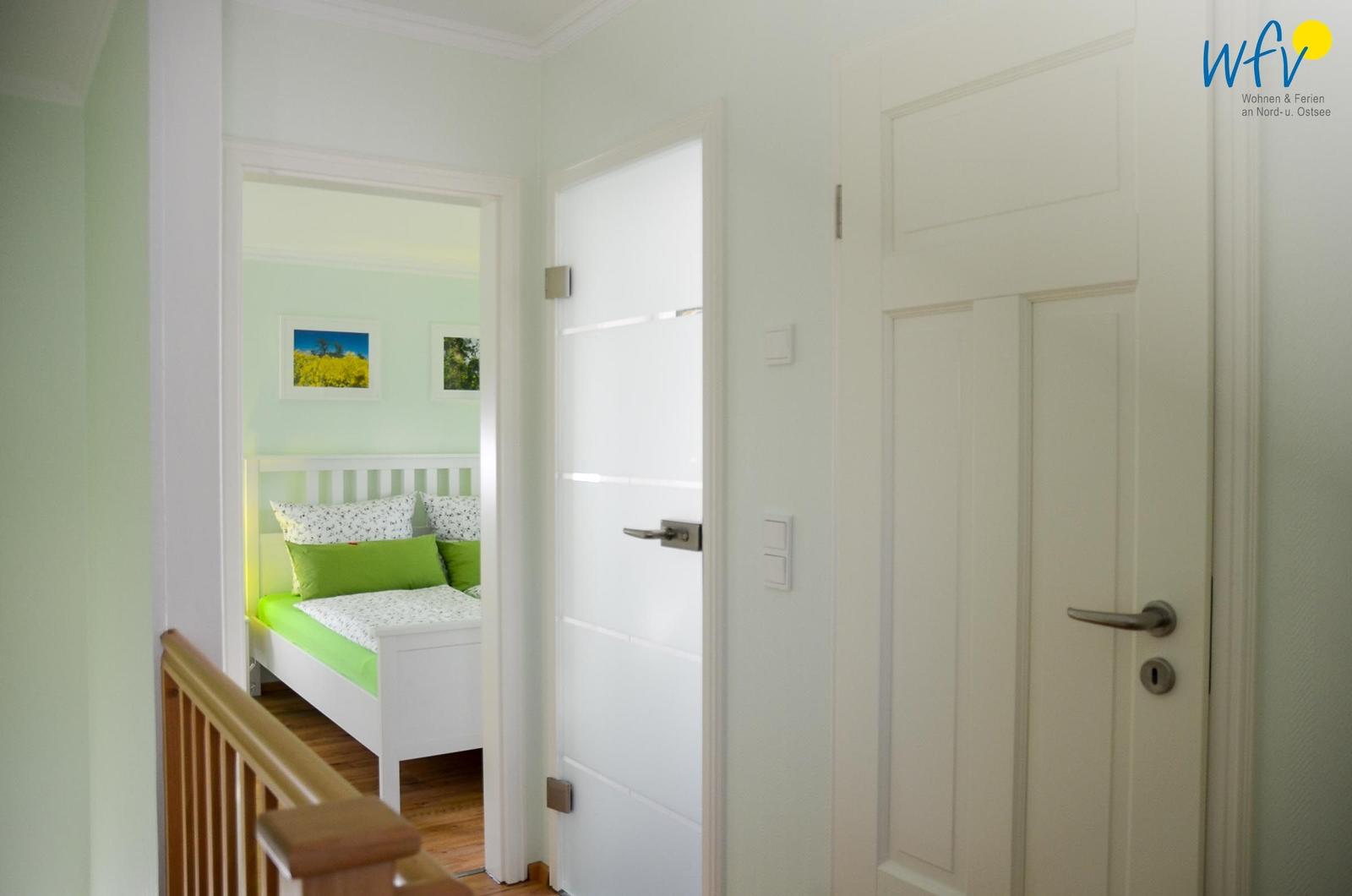 Schlafzimmer mit weißem Bett und grüner Bettwäsche. Holzgeländer und weiße Türen im Flur.