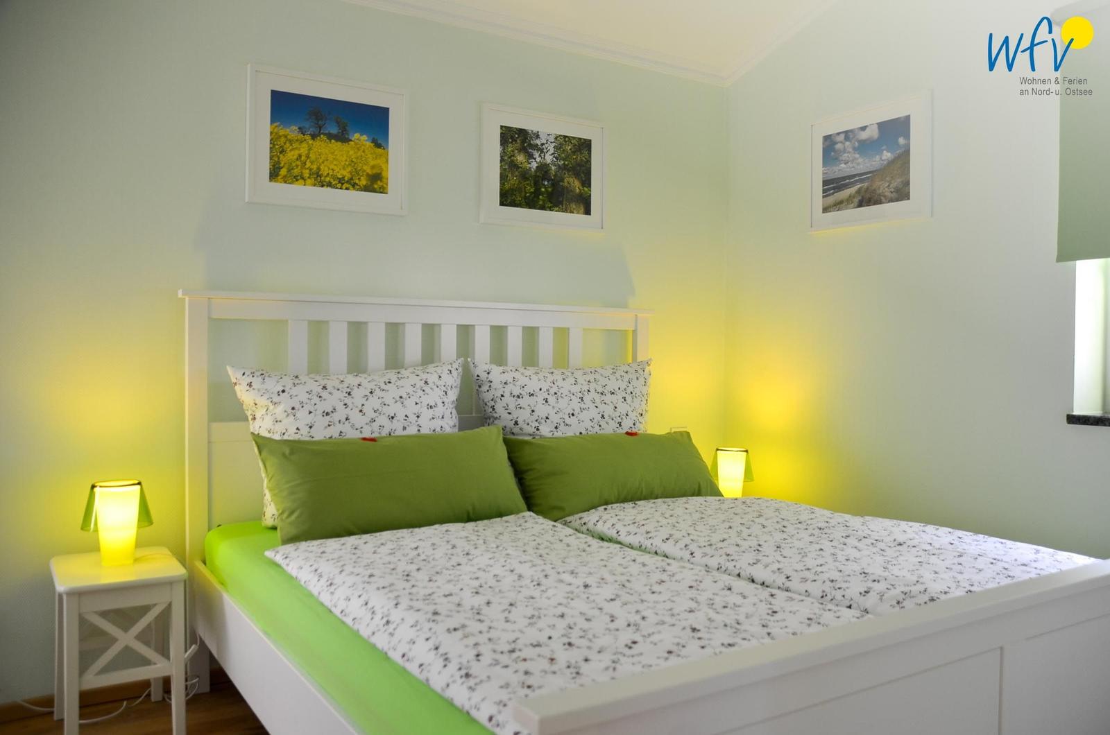 Schlafzimmer mit weißem Bett, grünen Kissen und Wandbildern.