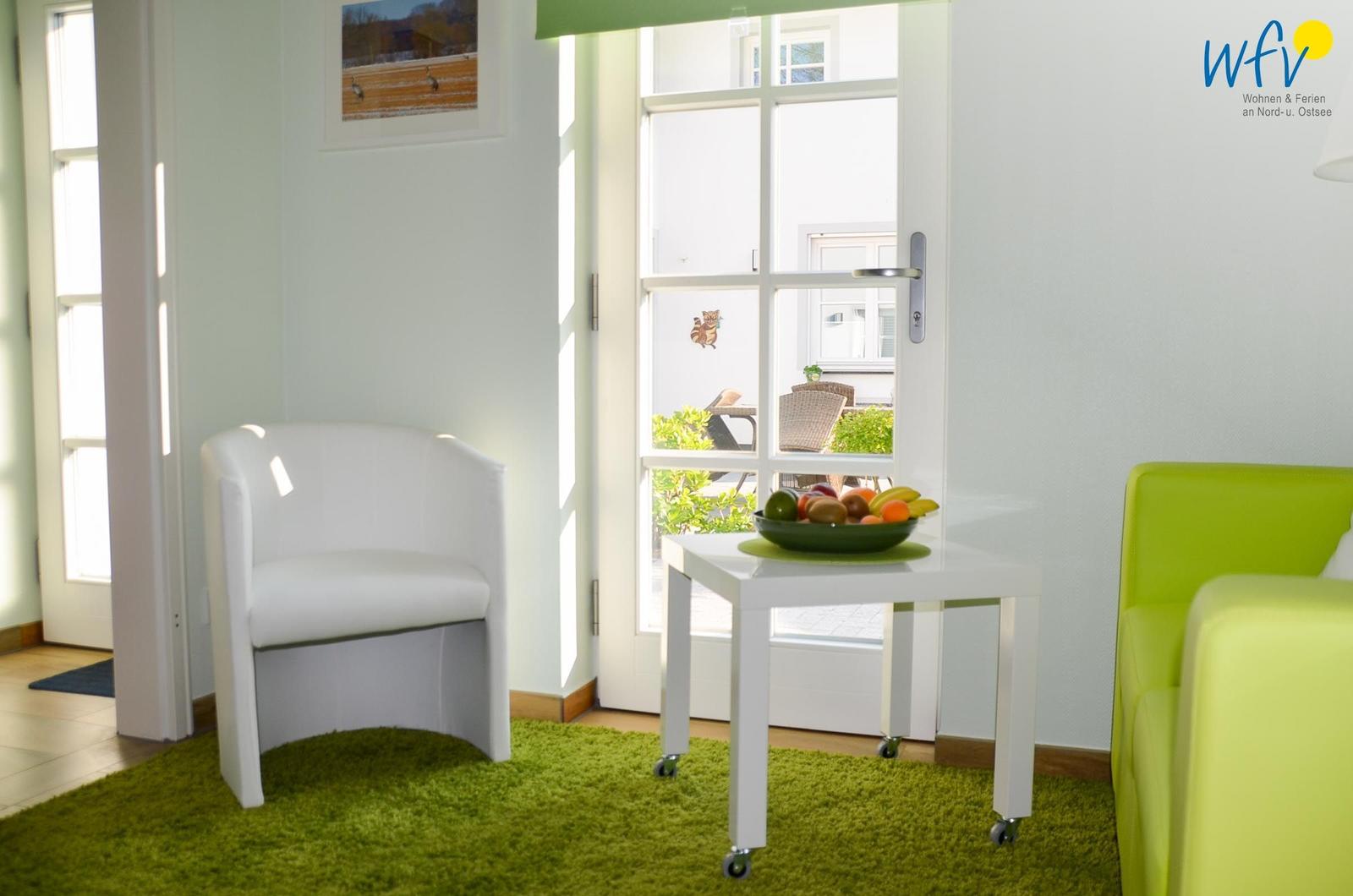 Ein Raum mit weißer Sitzgelegenheit, grünem Teppich und Tisch mit Obst. Durchgezogene Tür zum Außenbereich.