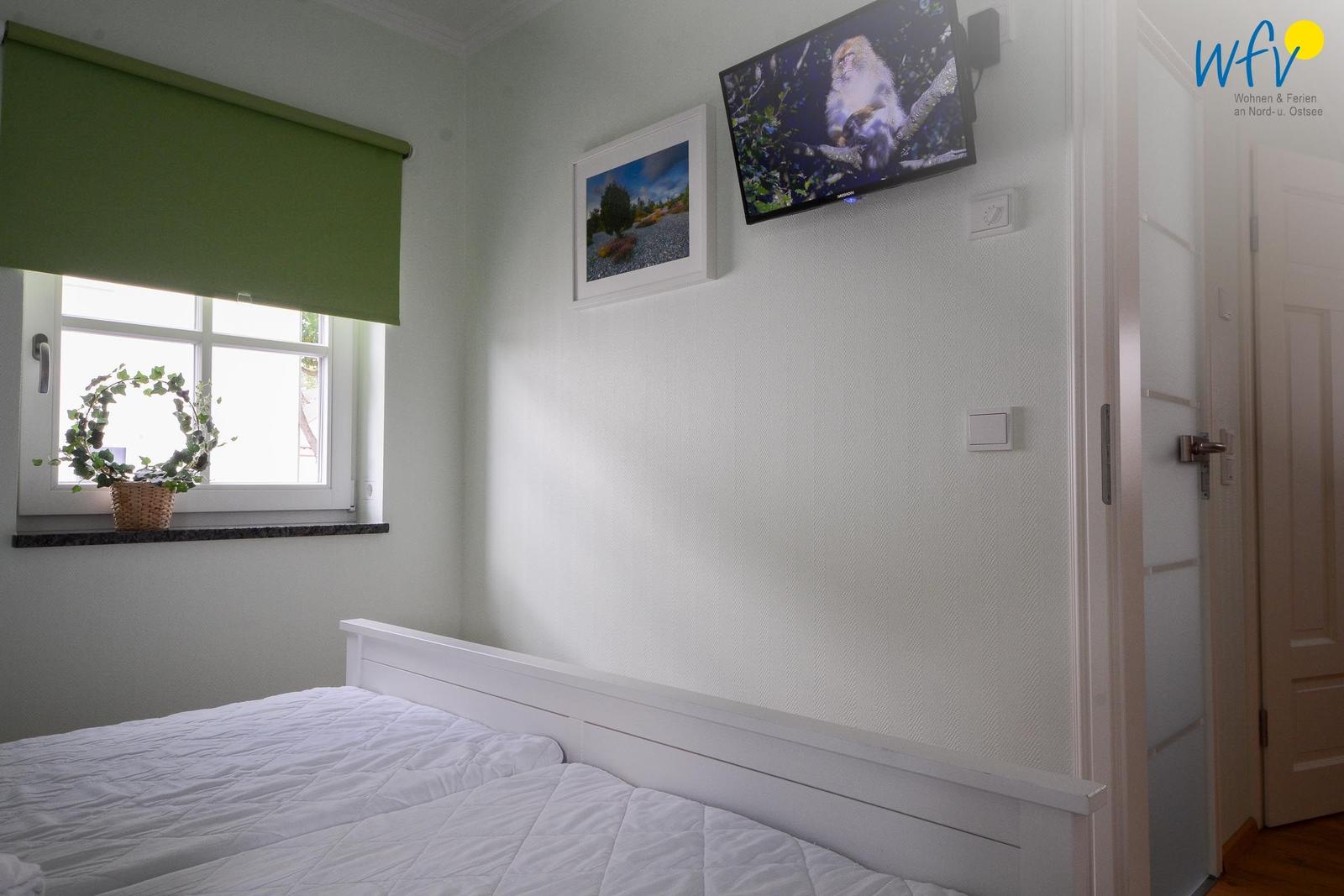 Ein Schlafzimmer mit weißem Bett, Wand-TV und Fenster mit grünem Rollladen.