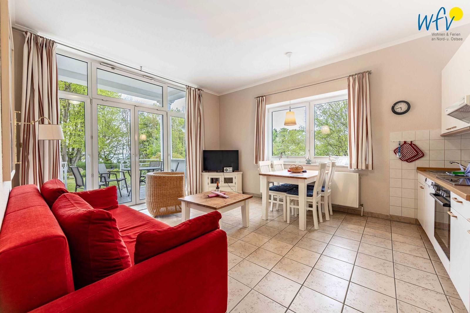 Wohnzimmer mit rotem Sofa, Essbereich und Küche. Durchgänge zu Balkon mit Gartenblick.