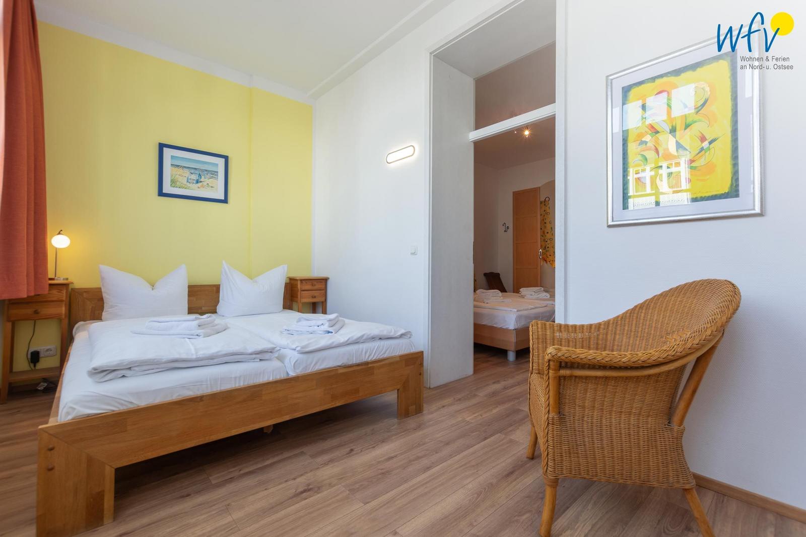 Schlafzimmer mit Doppelbett, Wickerstuhl und gelber Wand. Blick in angrenzende Zimmer.