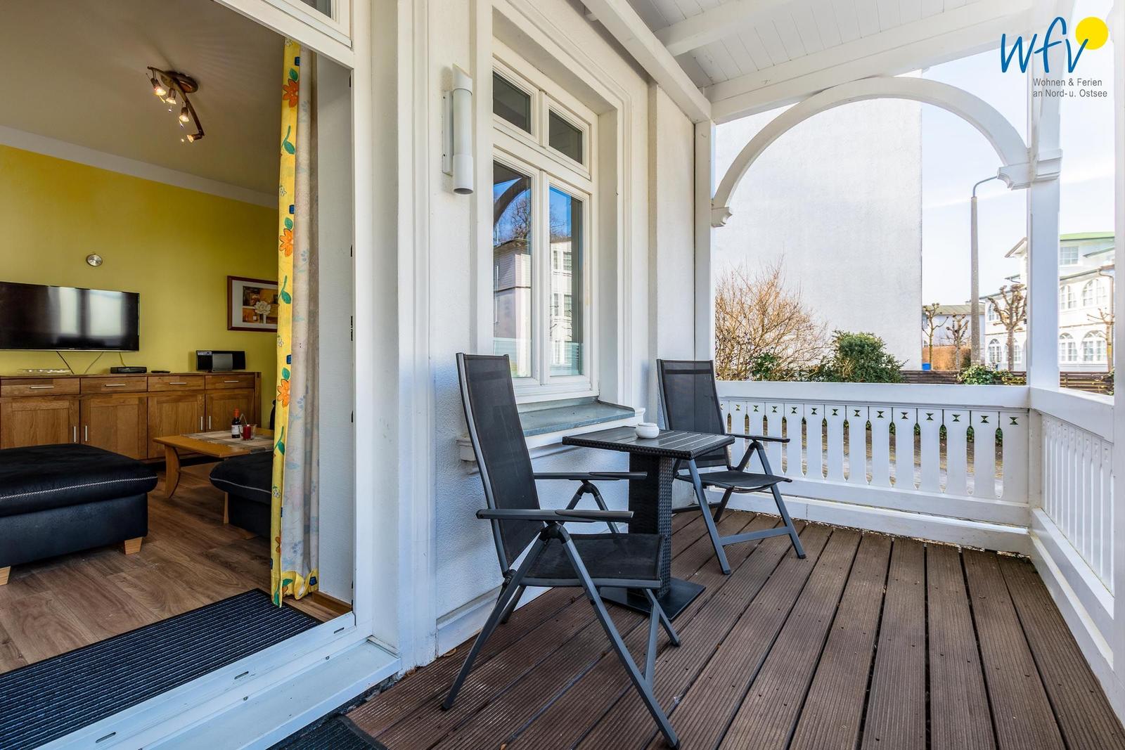 Terrasse mit Tisch und zwei Stühlen. Blick in Wohnzimmer mit Fernseher und Holzmöbeln.