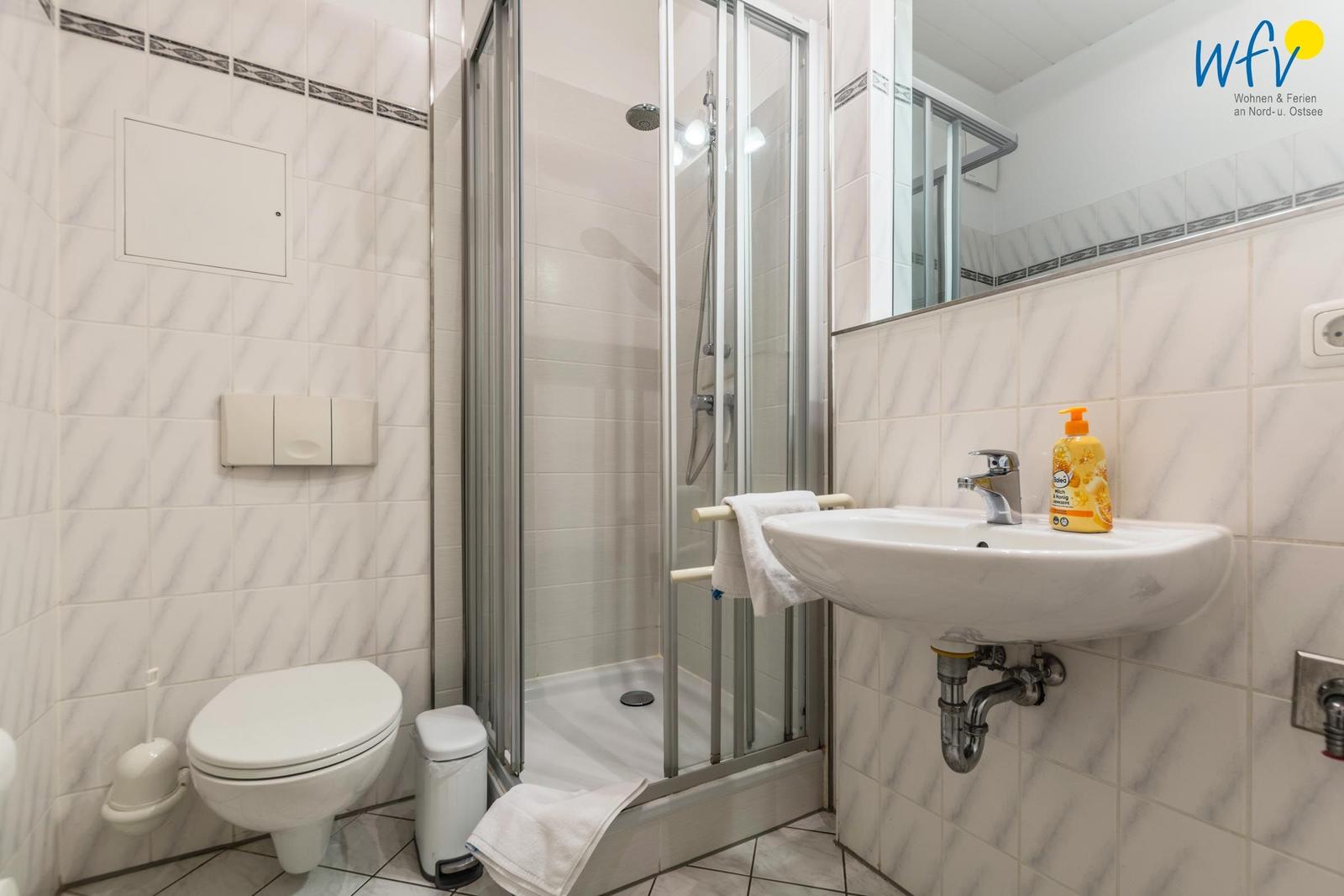 Badezimmer mit Dusche, Waschbecken und Toilette. Fliesenwand und Spiegel sichtbar.