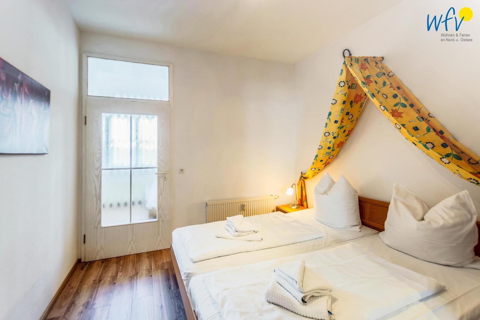 Schlafzimmer mit Doppelbett, gelbem Baldachin und Holzboden. Tür mit Glasfenster.