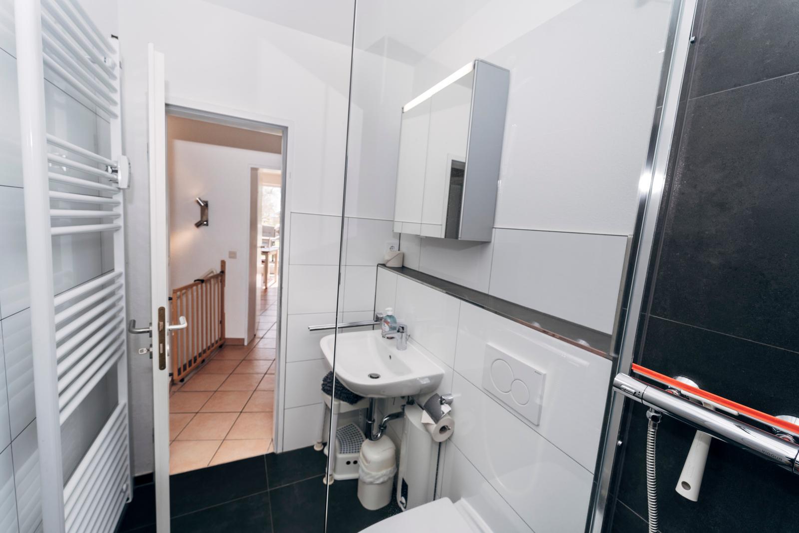 Badezimmer mit Dusche, Waschbecken und Heizkörper. Tür führt in Wohnbereich.